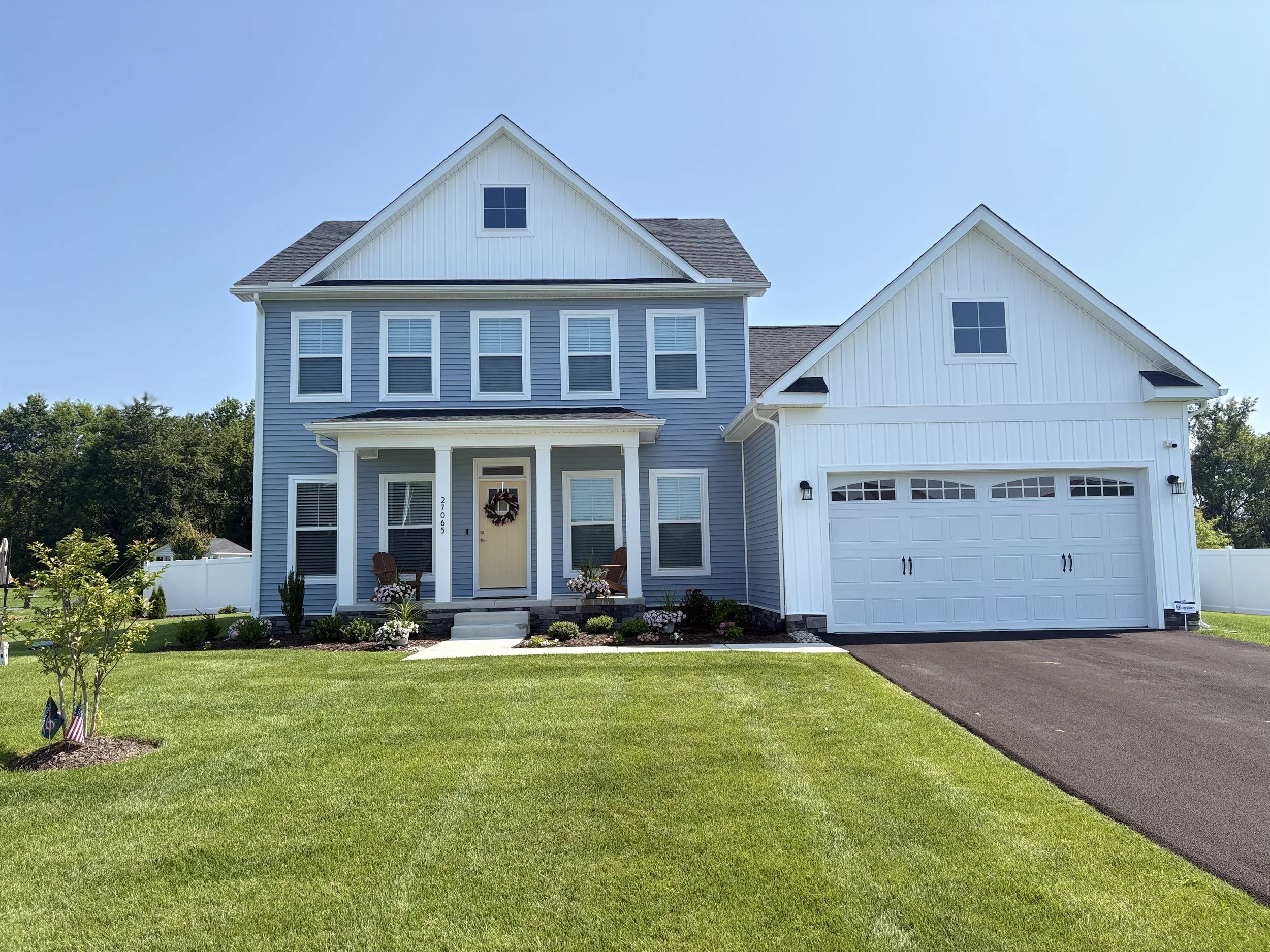 A modern two-story house with blue siding and white trim, a front porch, and a two-car garage. The house has a decorative wreath on the front door and a manicured lawn with small trees and flags.