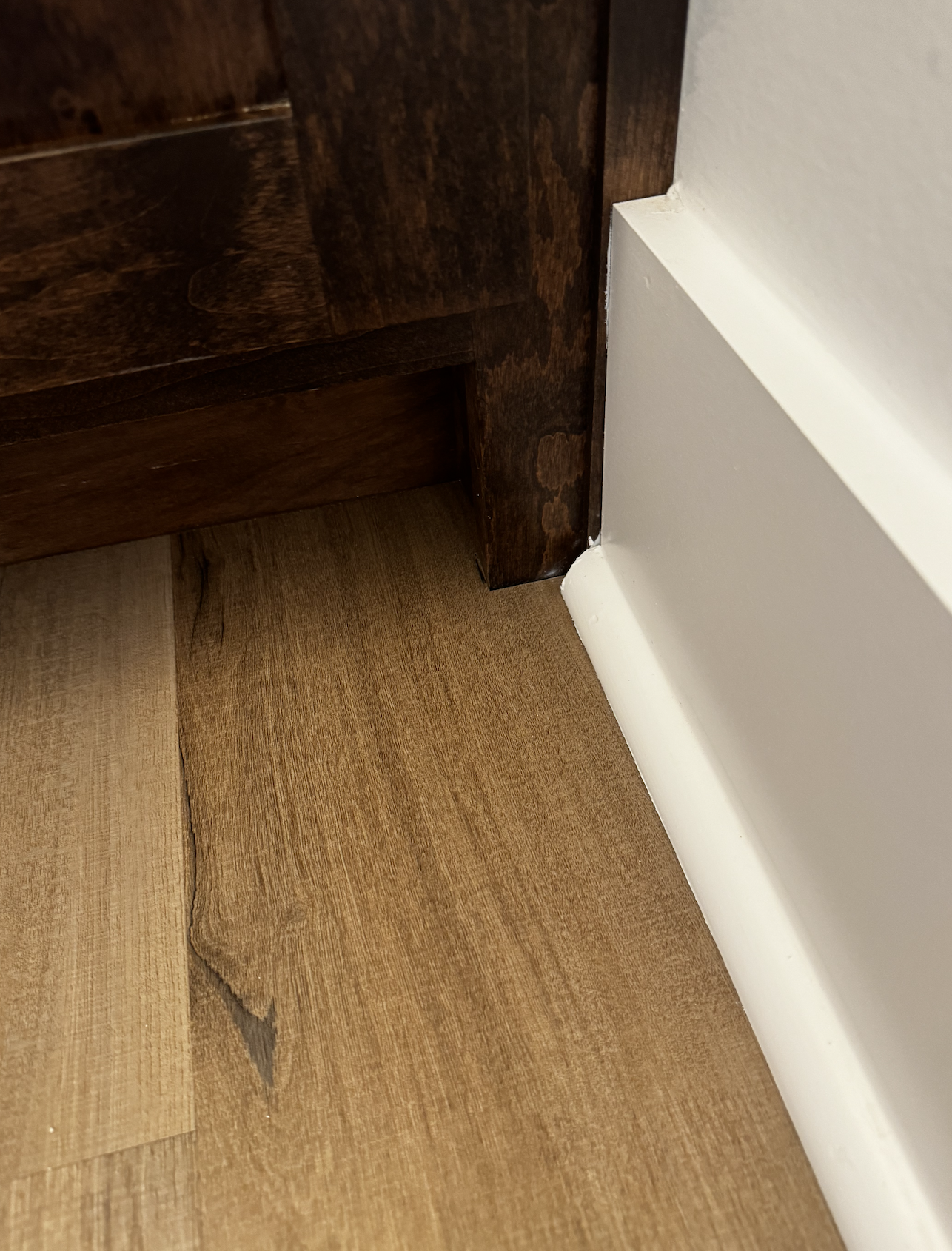 Close-up of a corner where a dark brown wooden cabinet meets a white wall and brown wooden flooring.