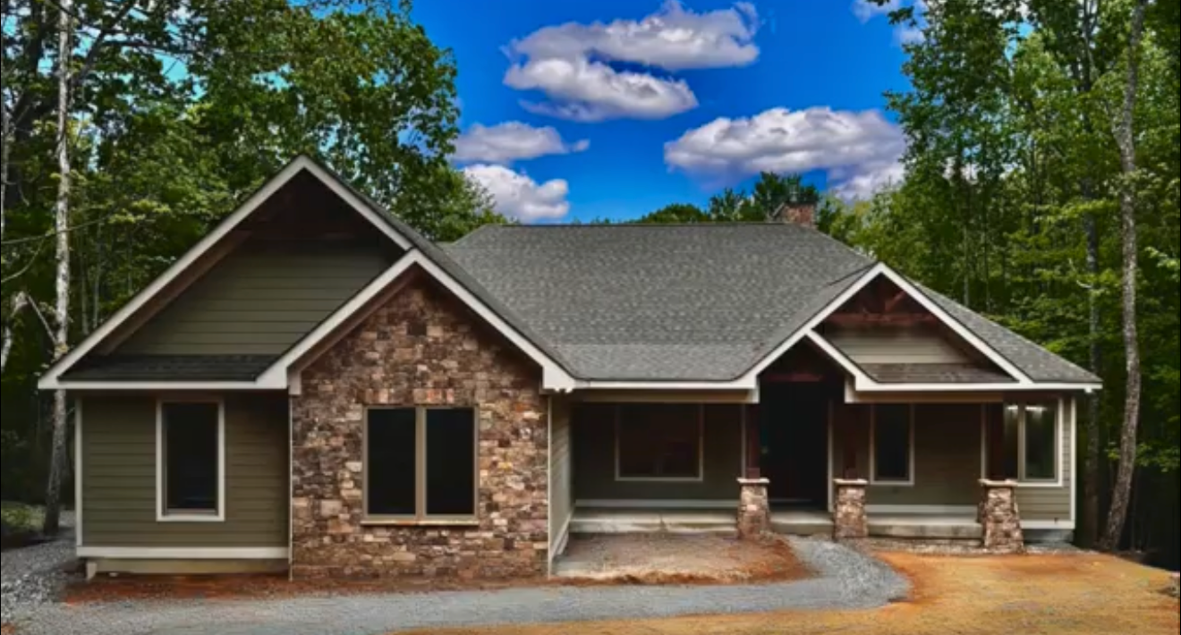 New house under construction with a gravel driveway, surrounded by trees, with a partly cloudy blue sky.
