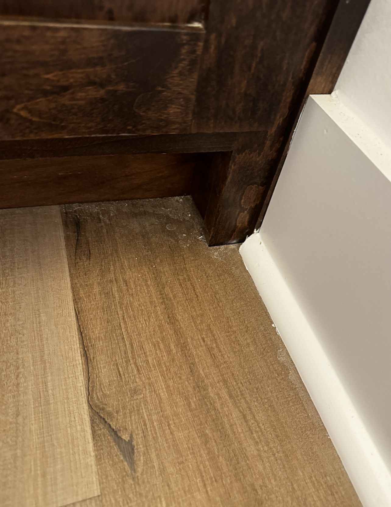 Close-up of a wooden piece of furniture meeting a white wall and hardwood flooring, showing some dust and dirt at the joint.