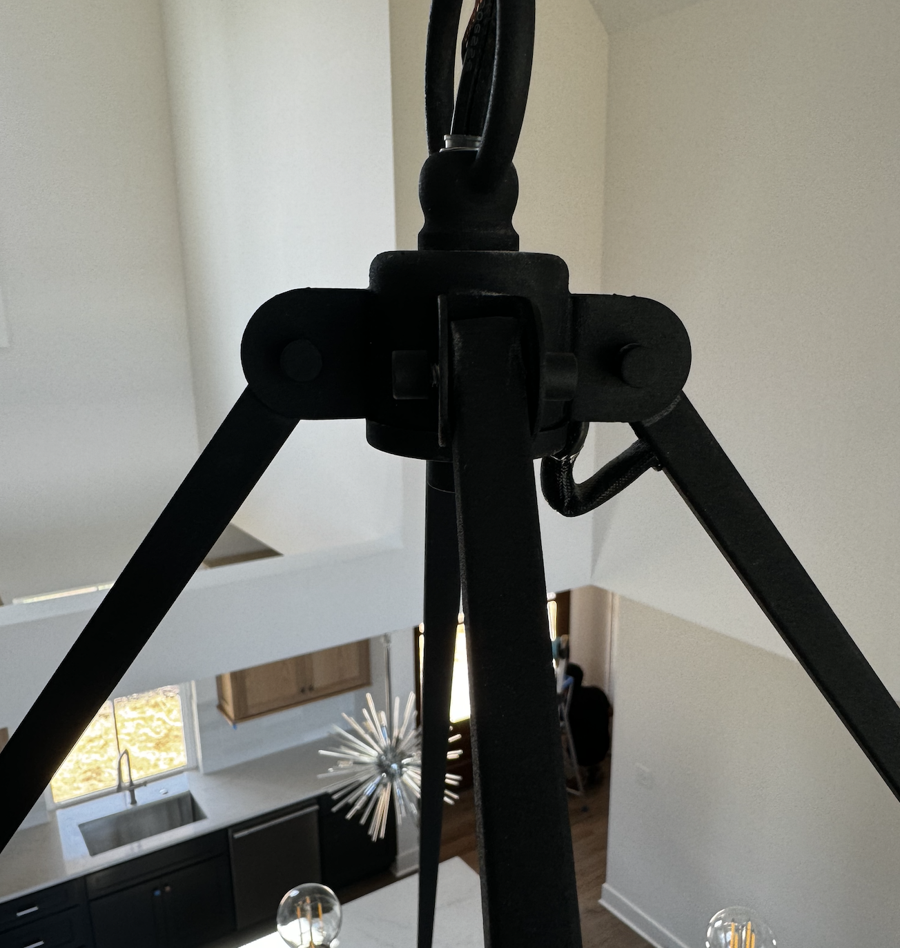 Close-up of a black metal tripod mount in a home interior, with a kitchen and living room visible in the background.