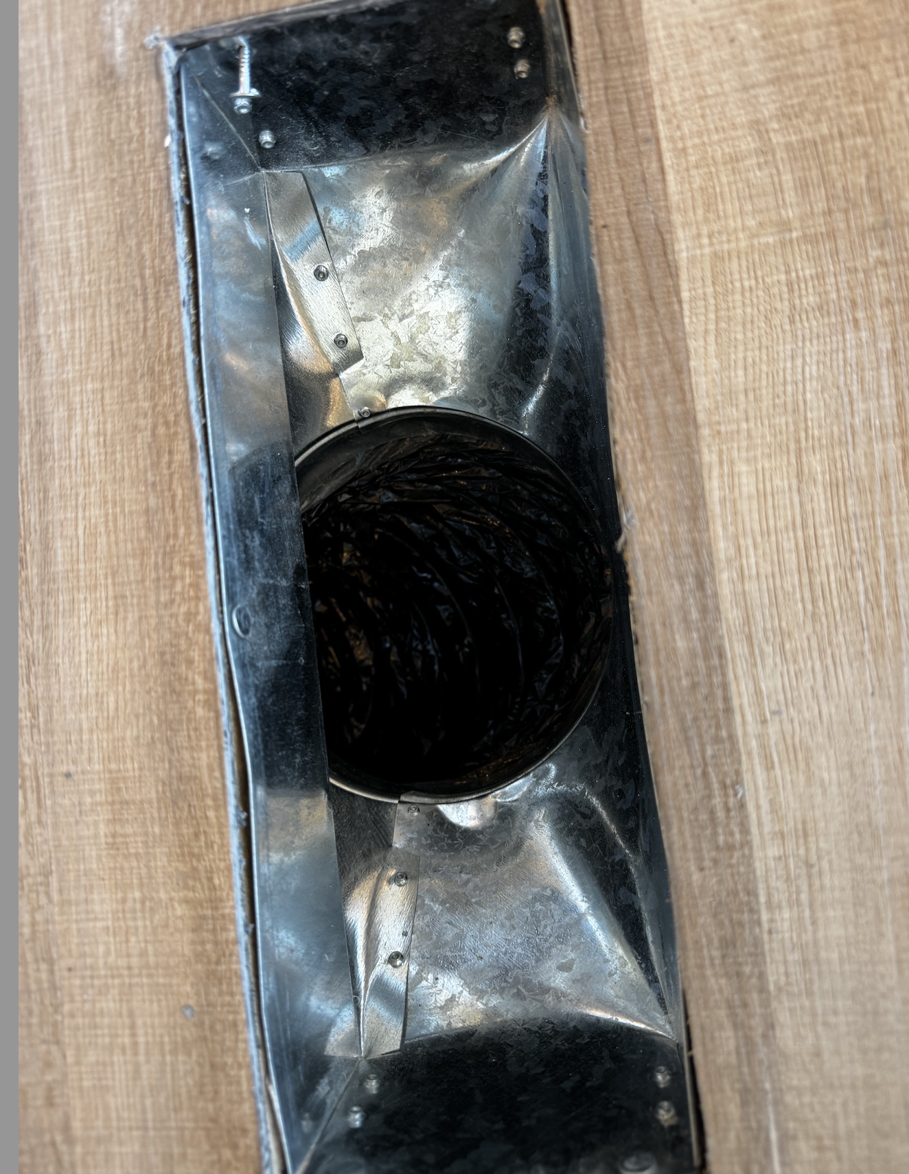Metal duct vent installed in wooden surface, black interior.