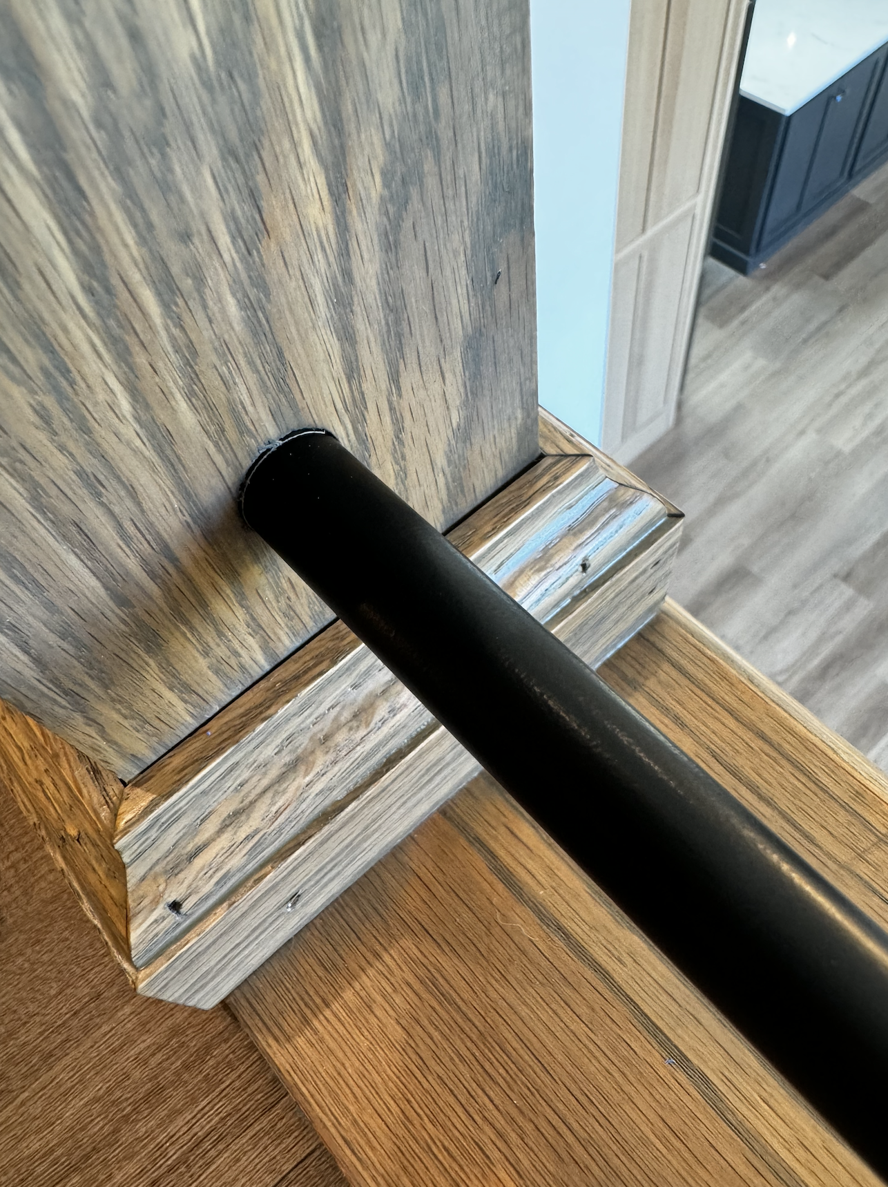 Close-up view of a black metal bar attached to a wooden post with a detailed wood grain finish in a room with wood flooring and cabinetry.