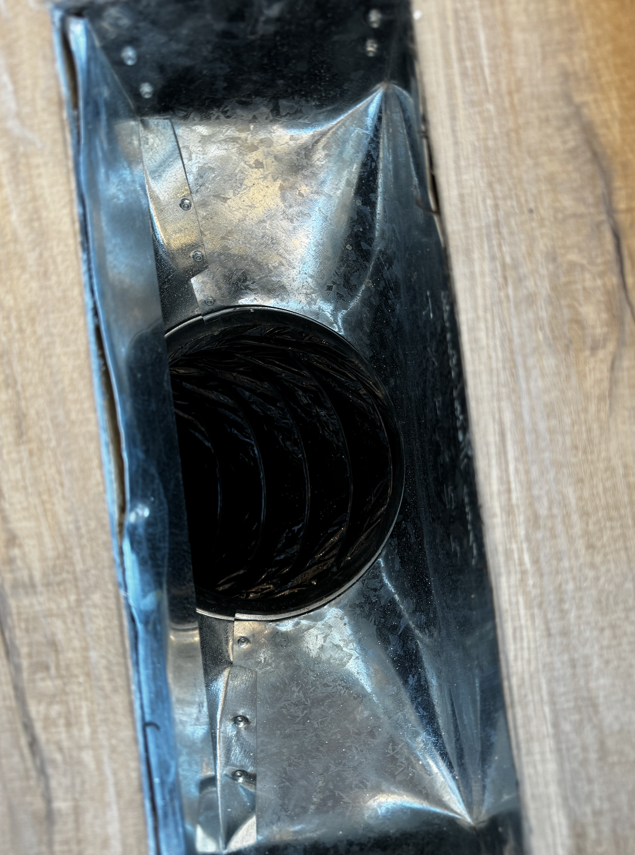 A metal vent or air duct with a black accordion-style flexible hose attached, installed in a wooden surface.
