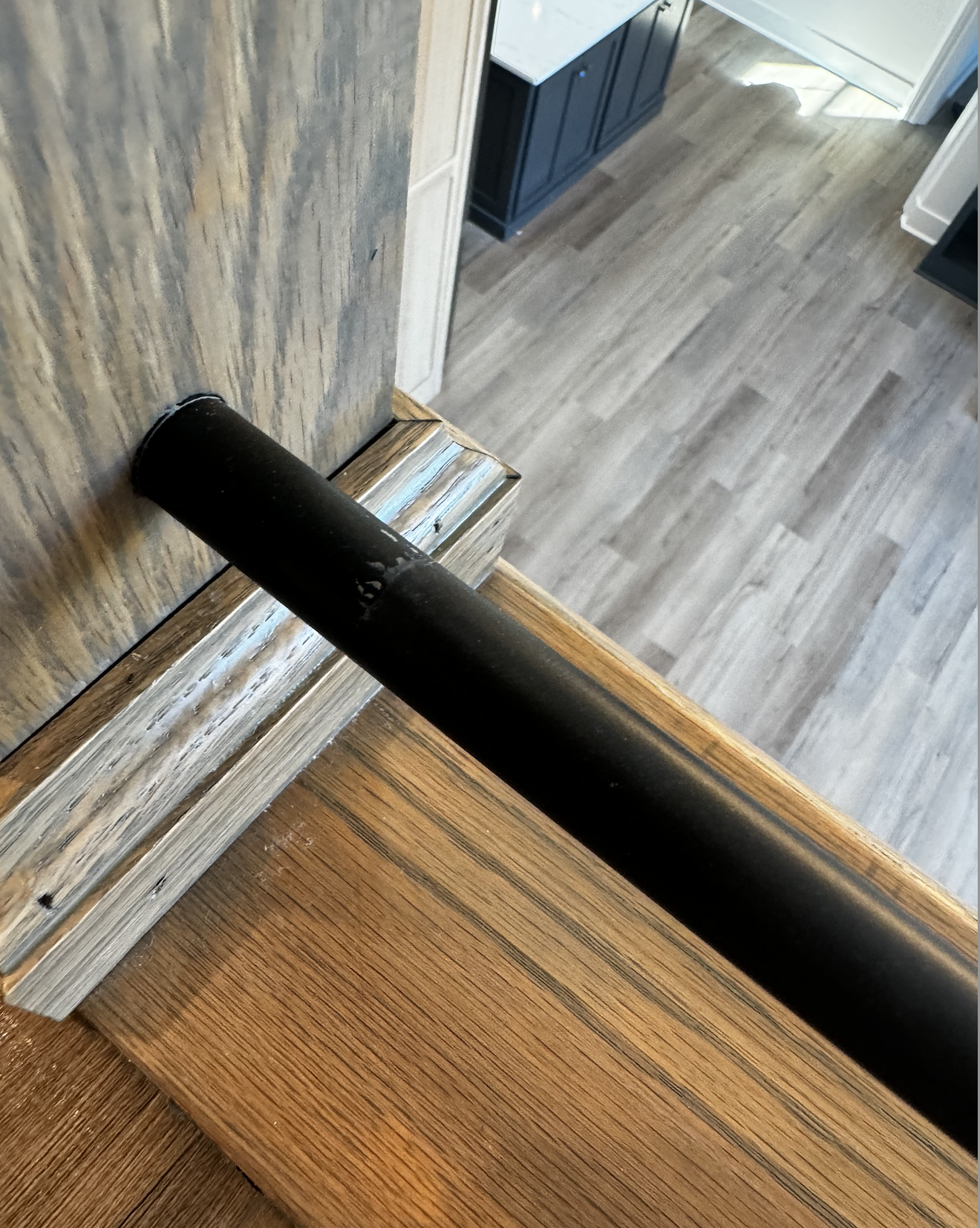 Close-up of a black handrail on a staircase with wooden steps, looking down towards a floor with grayish wood flooring and a dark-colored cabinet in the background.