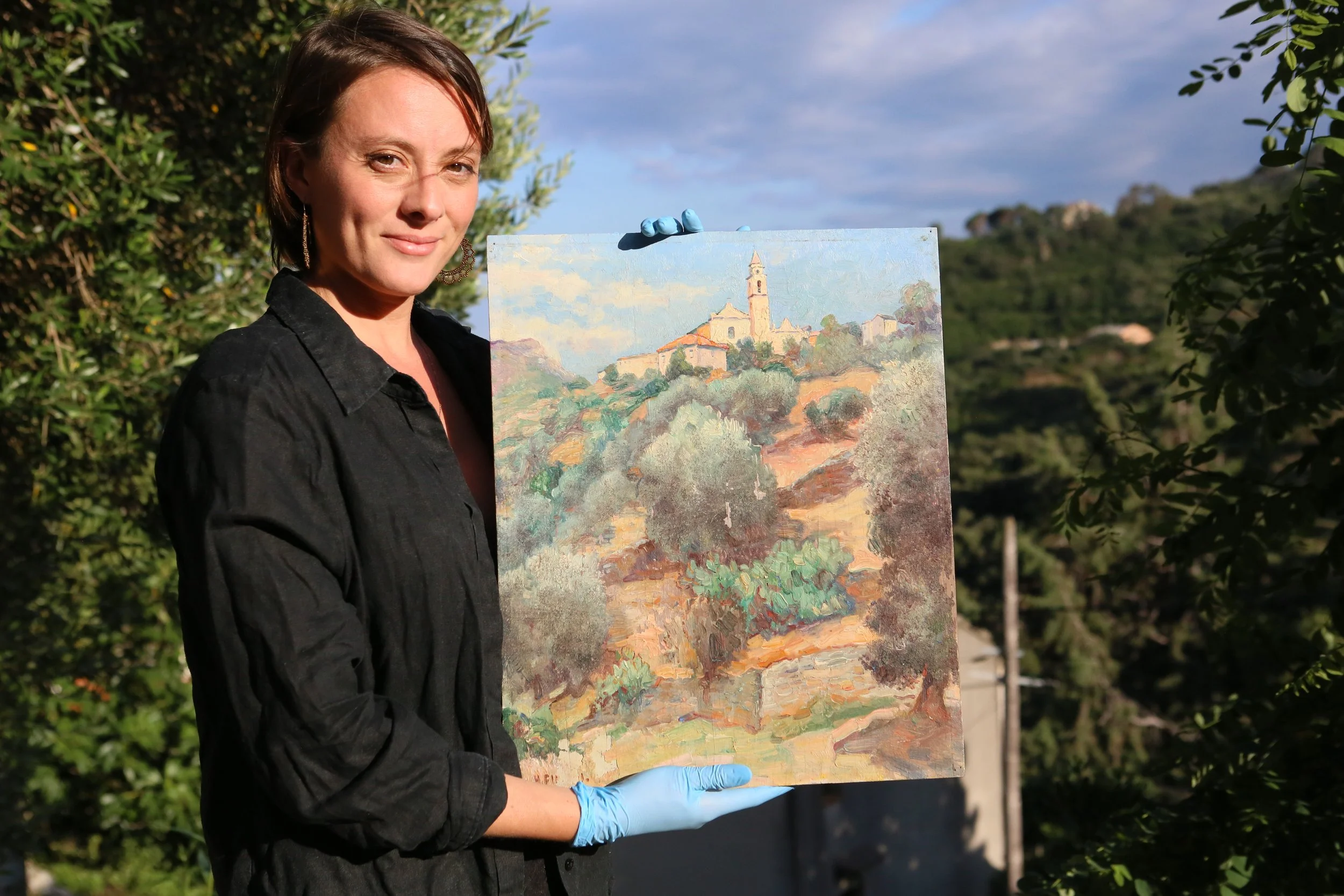 Femme tenant un tableau de peinture en plein air, dans un environnement naturel avec des arbres et un ciel bleu en arrière-plan.