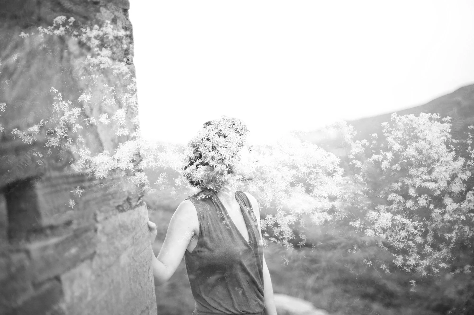 Une personne portant un t-shirt sans manches et un sac à dos, en plein air, avec des arbres et des montagnes en arrière-plan, entourée d'effets visuels de fleurs ou de feuillage en blanc et noir.