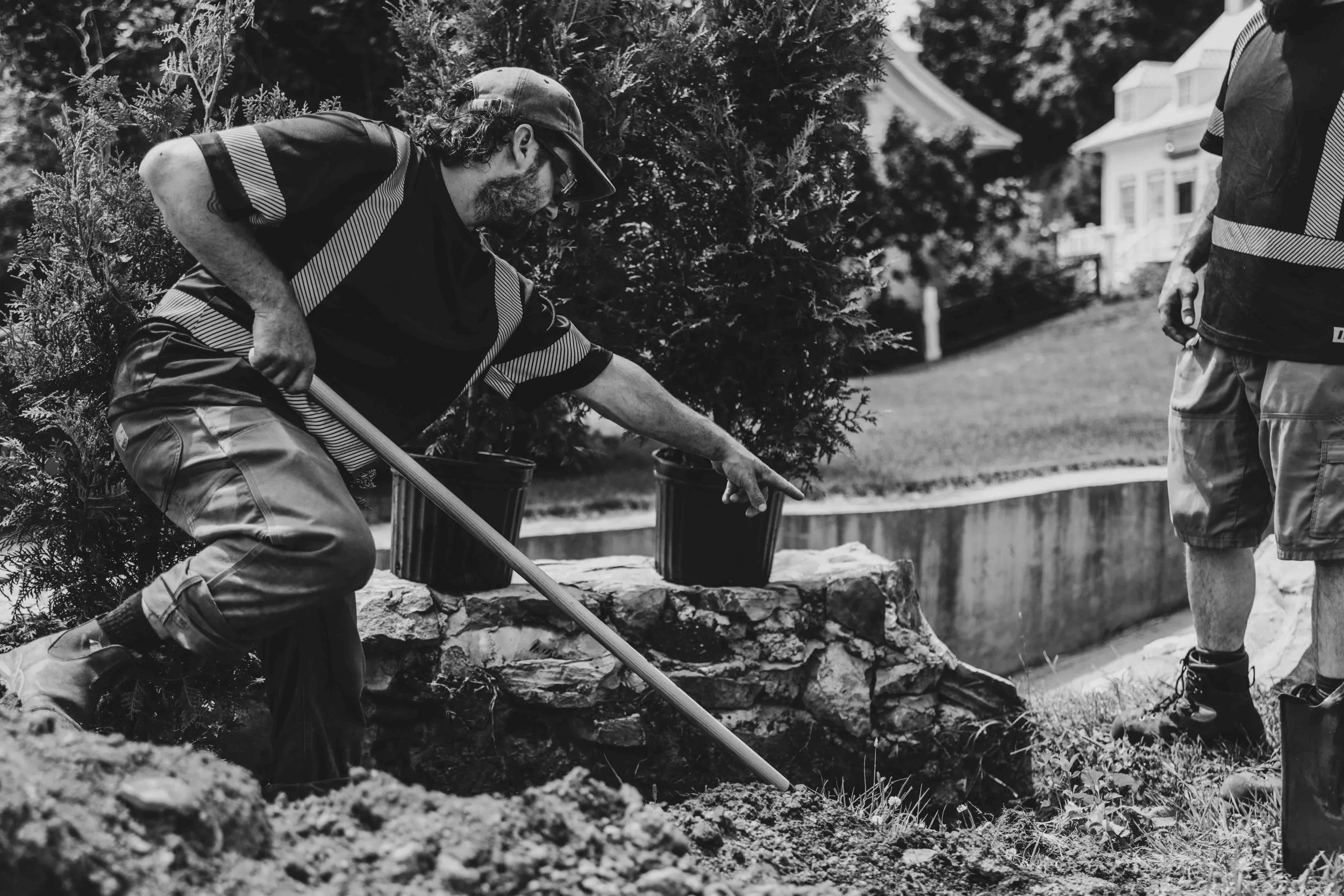 Deux hommes travaillant au jardin, l'un ramasse la terre avec une pelle tandis que l'autre se tient debout. Tous deux portent des vêtements décontractés, avec des t-shirts et des shorts, et un d'eux porte une casquette.