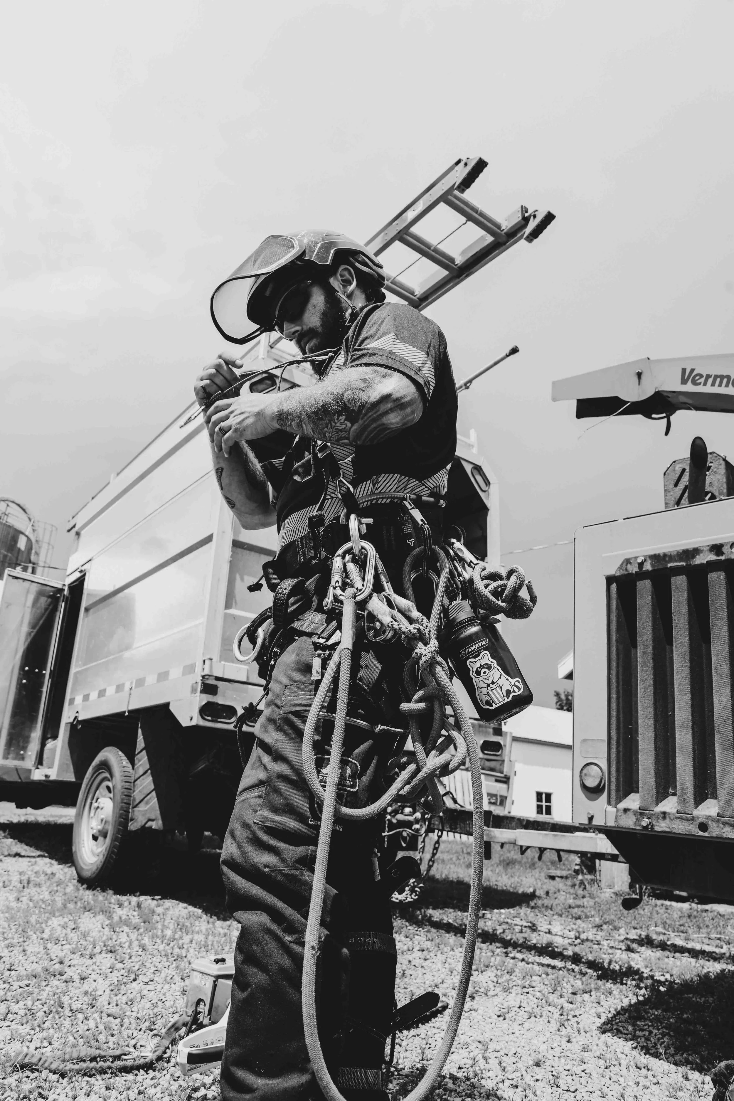 Un homme portant un casque de sécurité et un équipement de travail, ajustant une corde ou un câble, probablement en train de travailler en hauteur, avec un camion équipé de matériel de levage en arrière-plan.