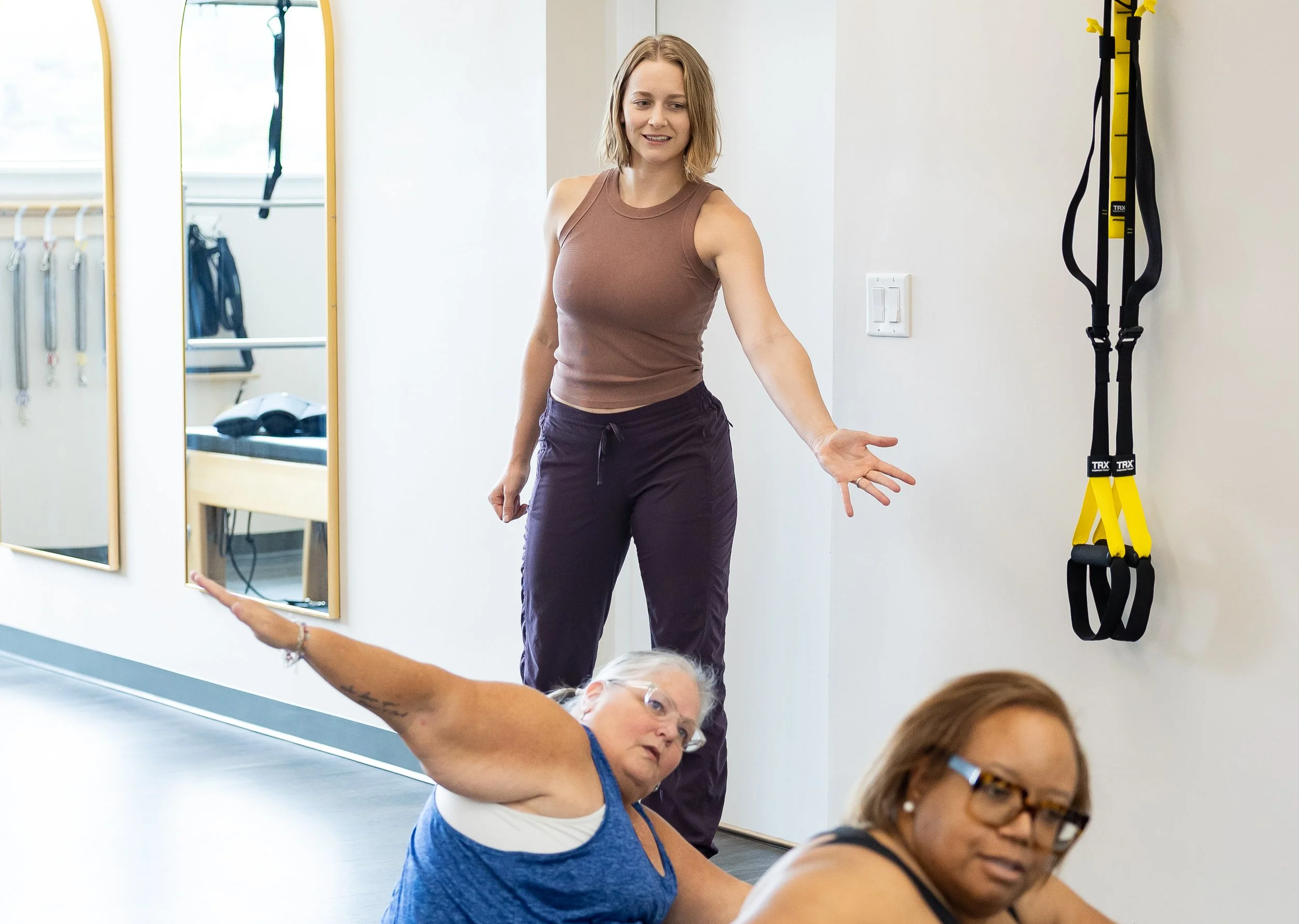 Instructor demonstrating a stretch with a client, Pilates equipment visible in the background.