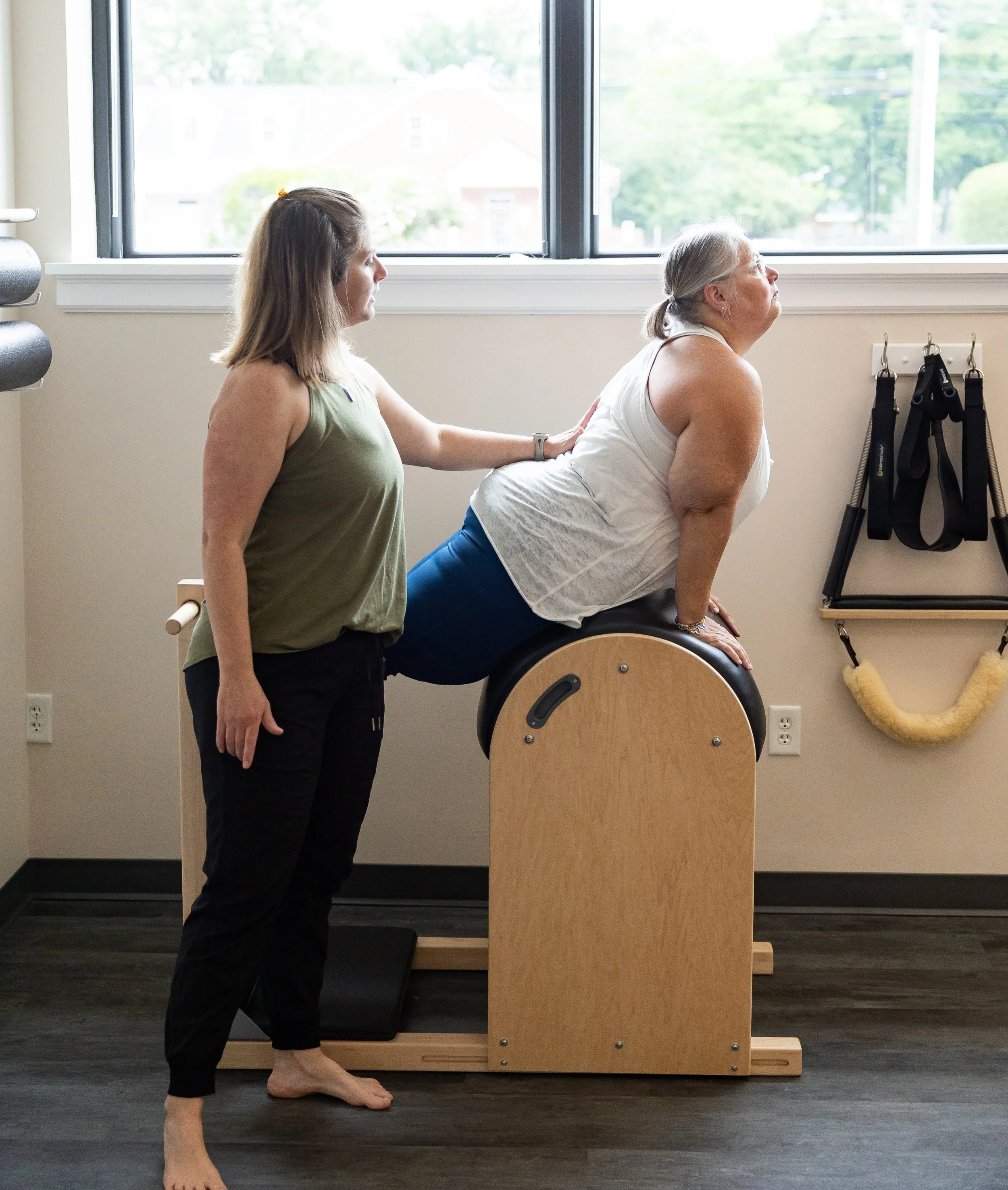 Client being assisted by an instructor on a Pilates chair, in the studio with large windows