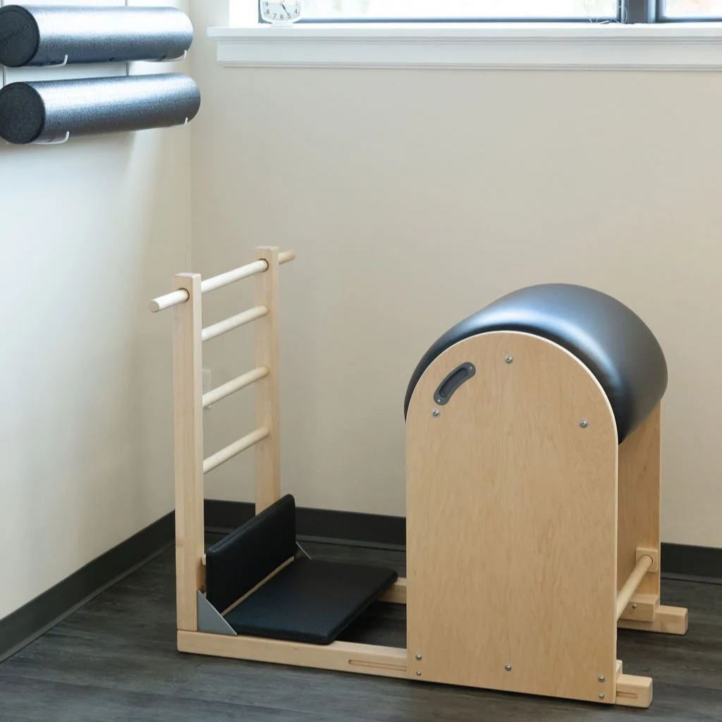 Pilates chair with padded footrest and handles, in a studio with a window, clock, and storage tubes on the wall