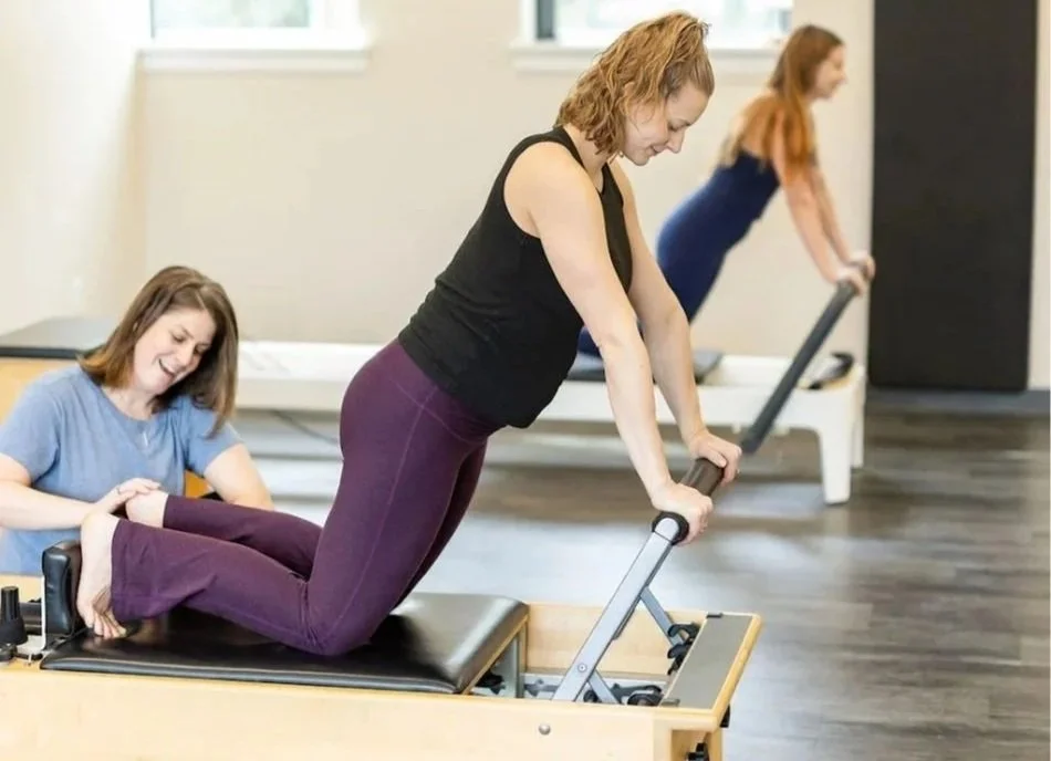 Instructor assisting a client with a leg stretch on a reformer, with another client exercising on a reformer in the background