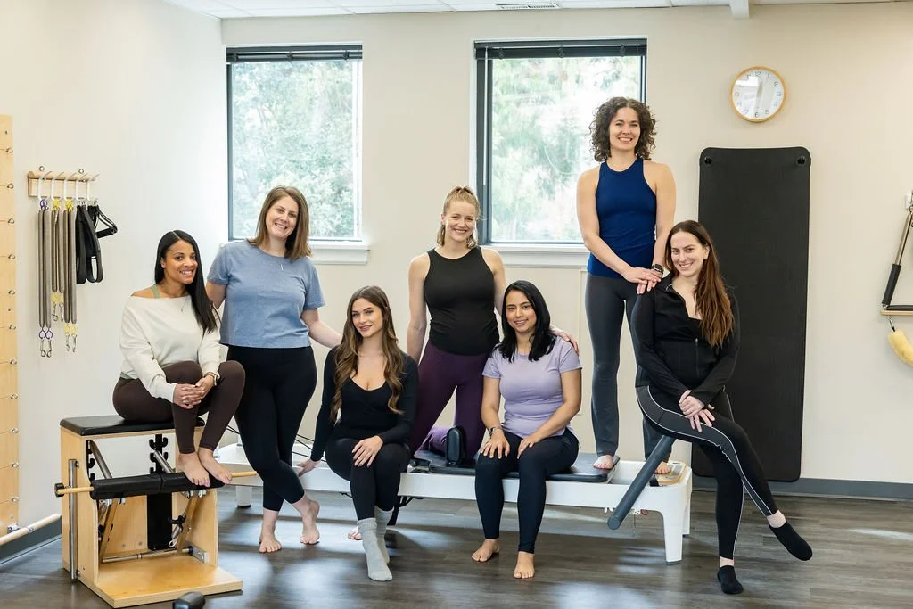 Pilates Plus instructor team smiling together in the studio, with Pilates equipment on the wall behind them.