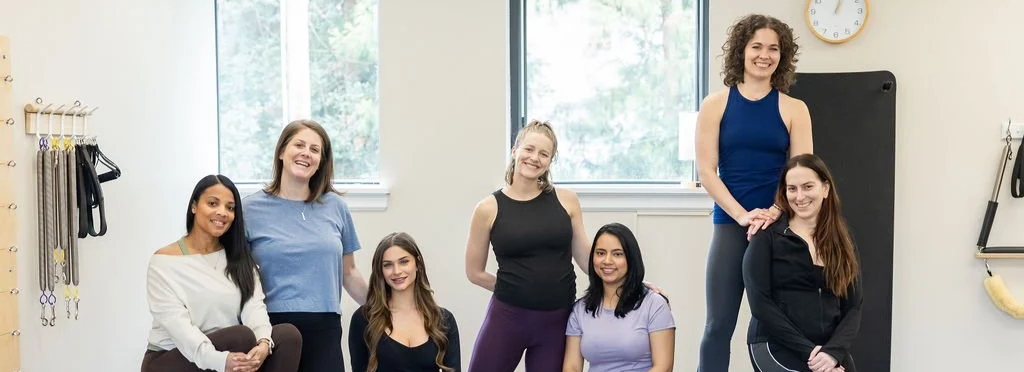 Pilates Plus instructor team smiling together in the studio, with Pilates equipment on the wall behind them