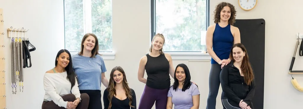 Pilates Plus instructor team smiling together in the studio, with Pilates equipment on the wall behind them