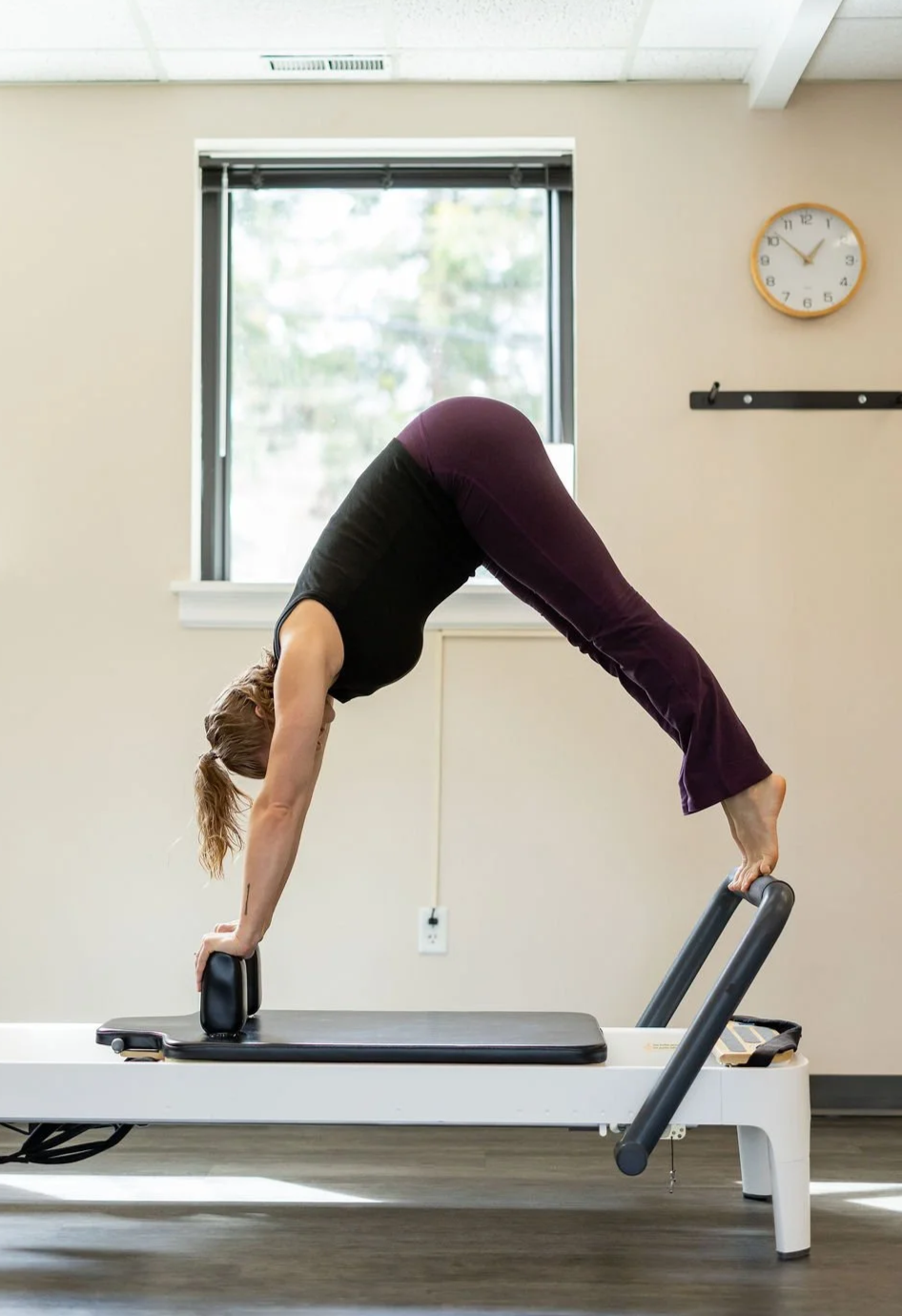 A woman practicing Pilates on a reformer machine, in a room with a window, clock, and wall hooks.