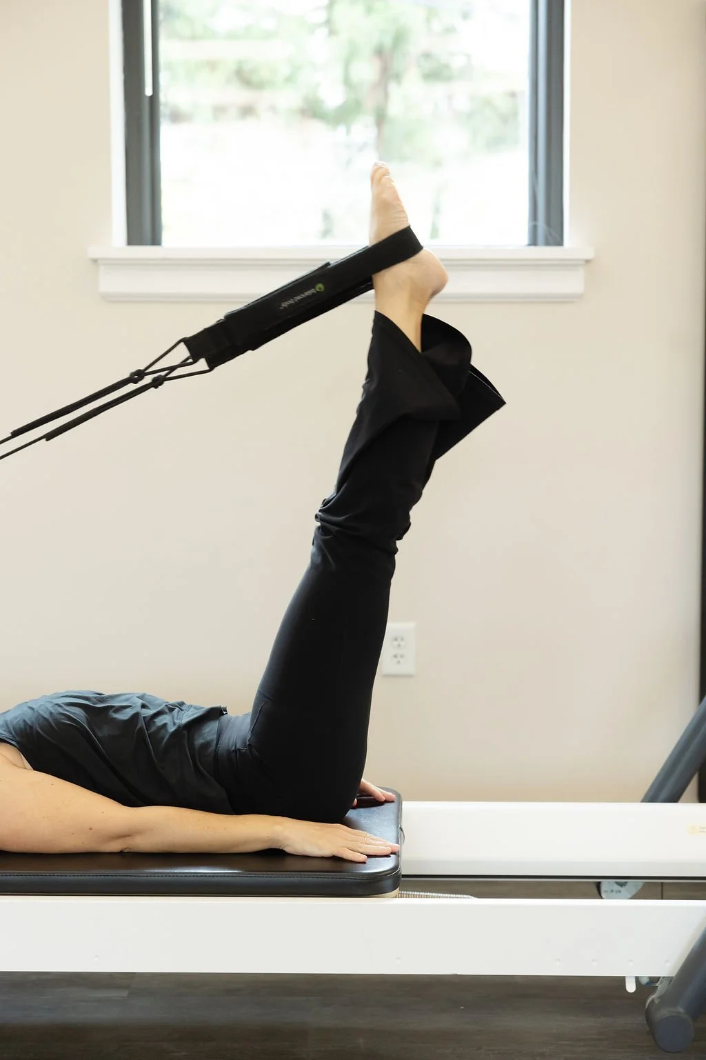 Client lying on her back on a reformer, performing leg work with one leg raised and strapped in