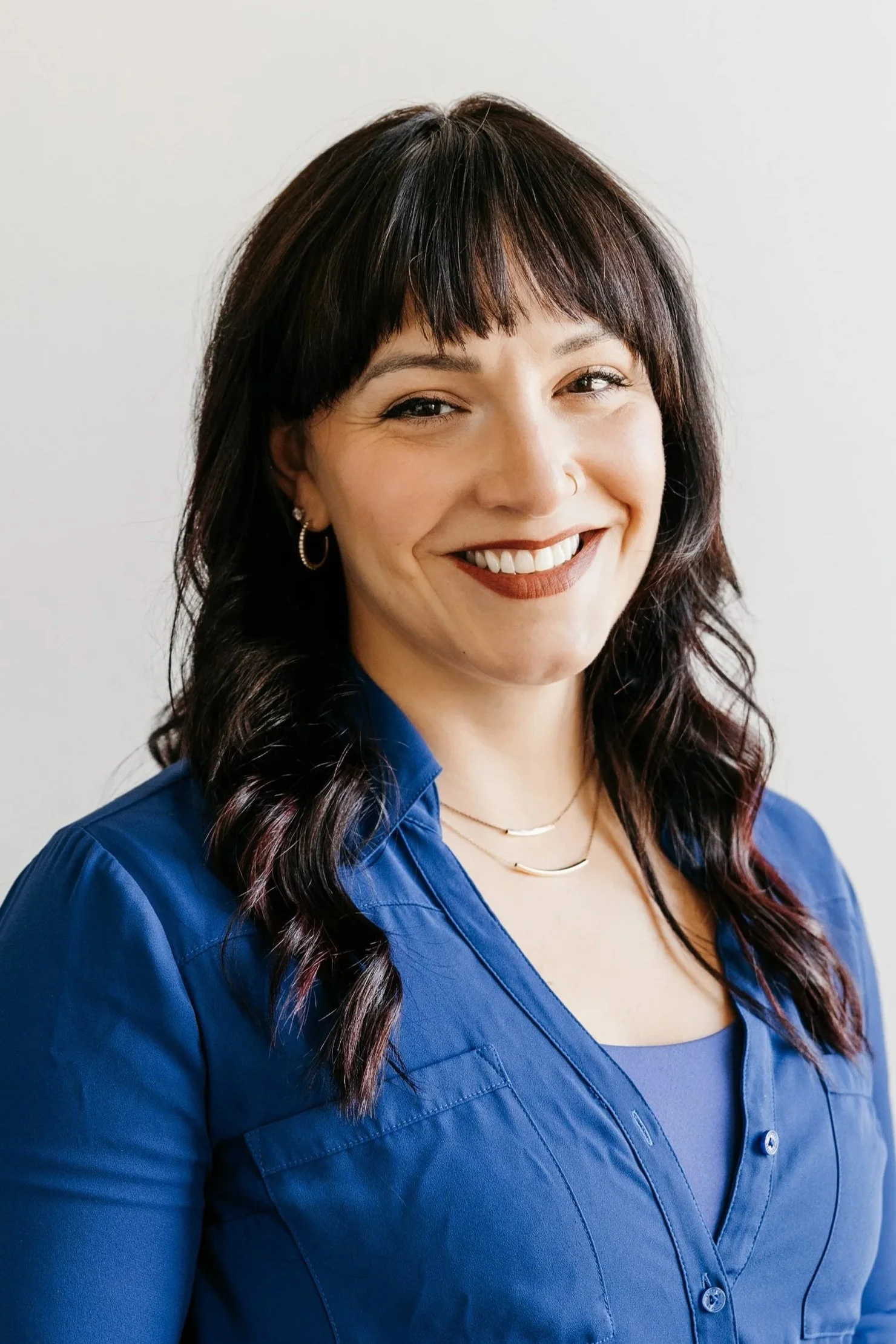 A smiling woman with dark hair and bangs, wearing a blue shirt and jewelry, standing against a plain white background.