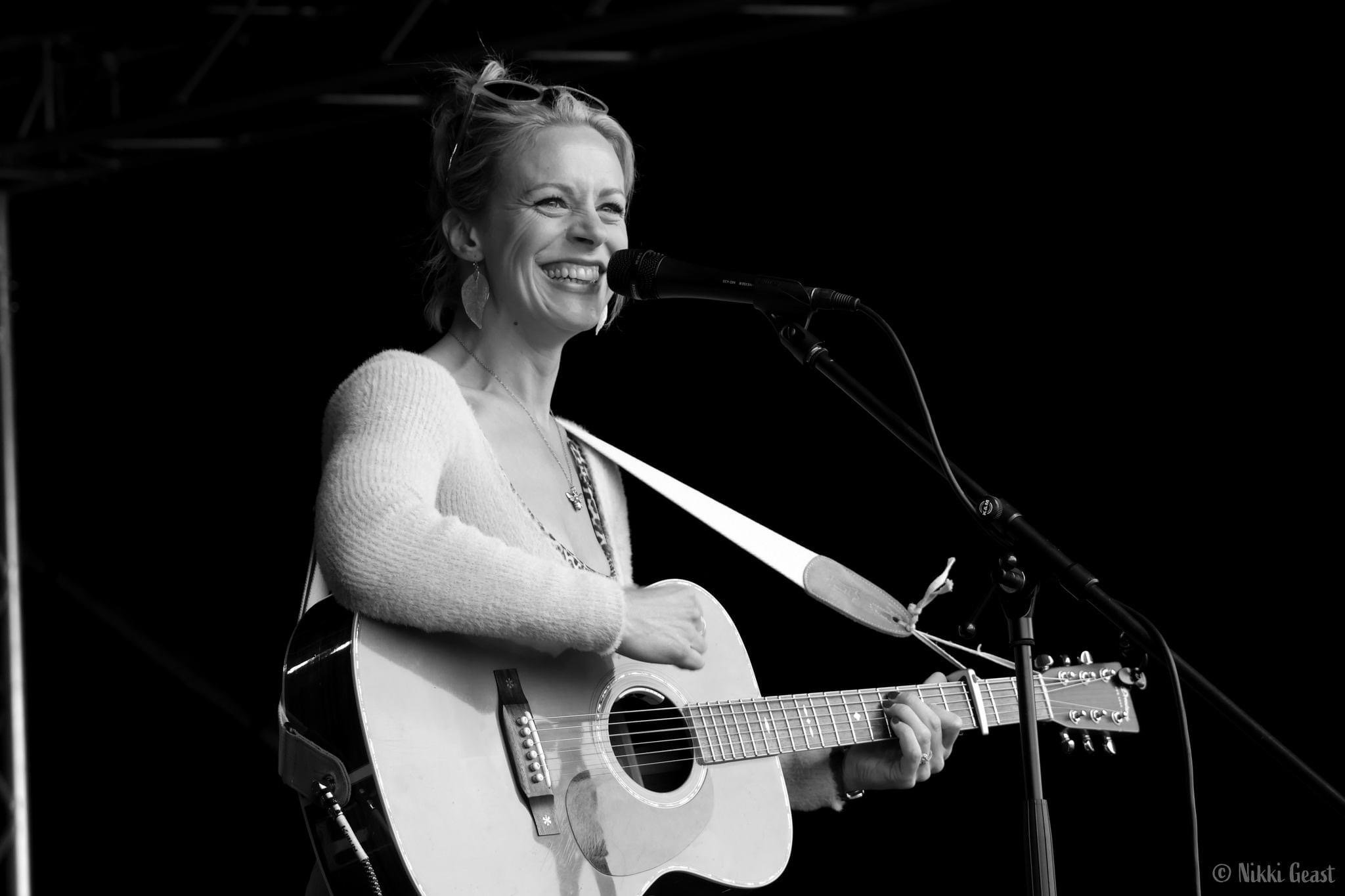 A woman smiling while playing an acoustic guitar and singing into a microphone on stage.