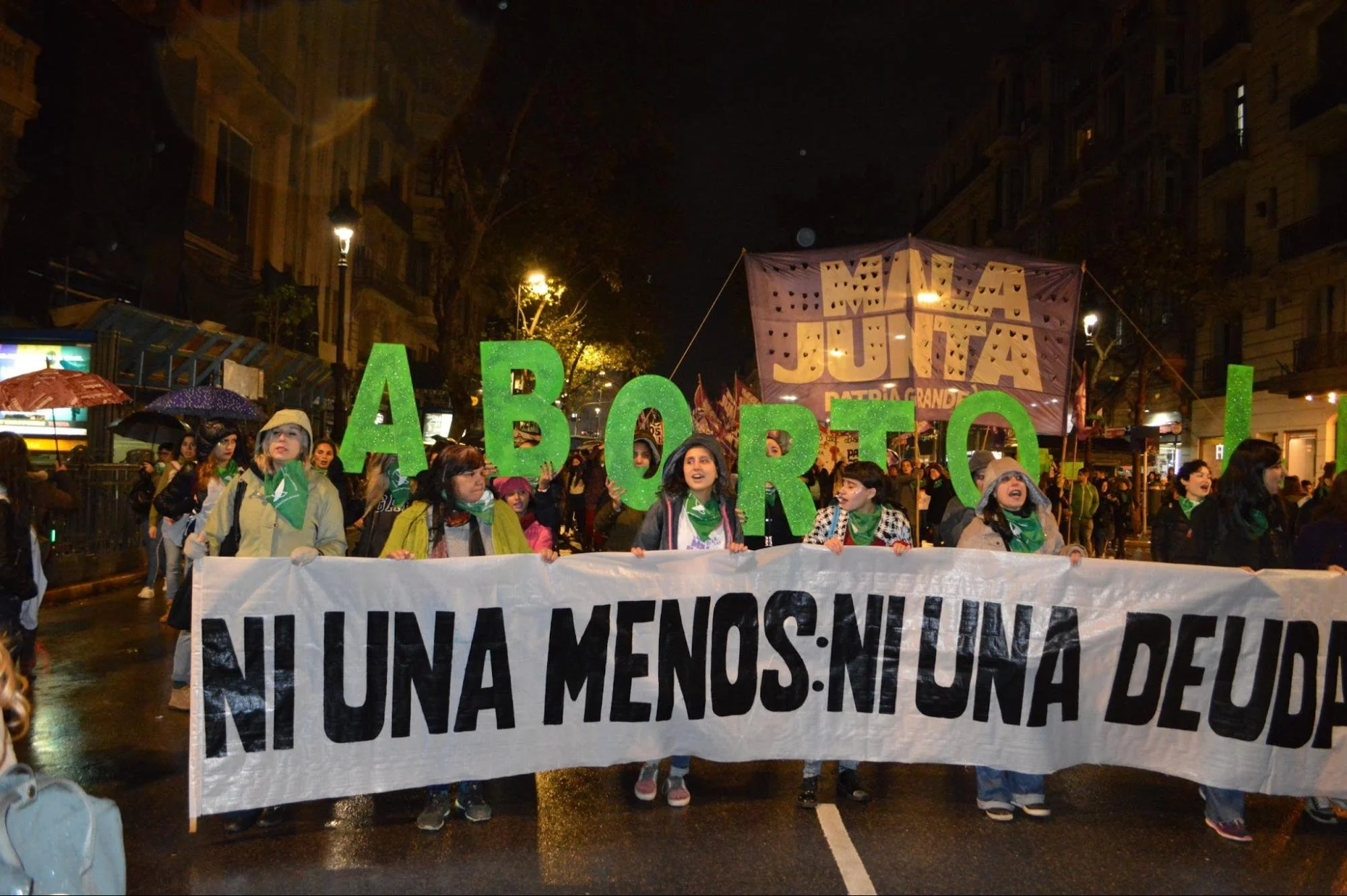Protesters marching at night holding umbrellas and banners, one reading 'Ni Una Menos', some wearing rain jackets.