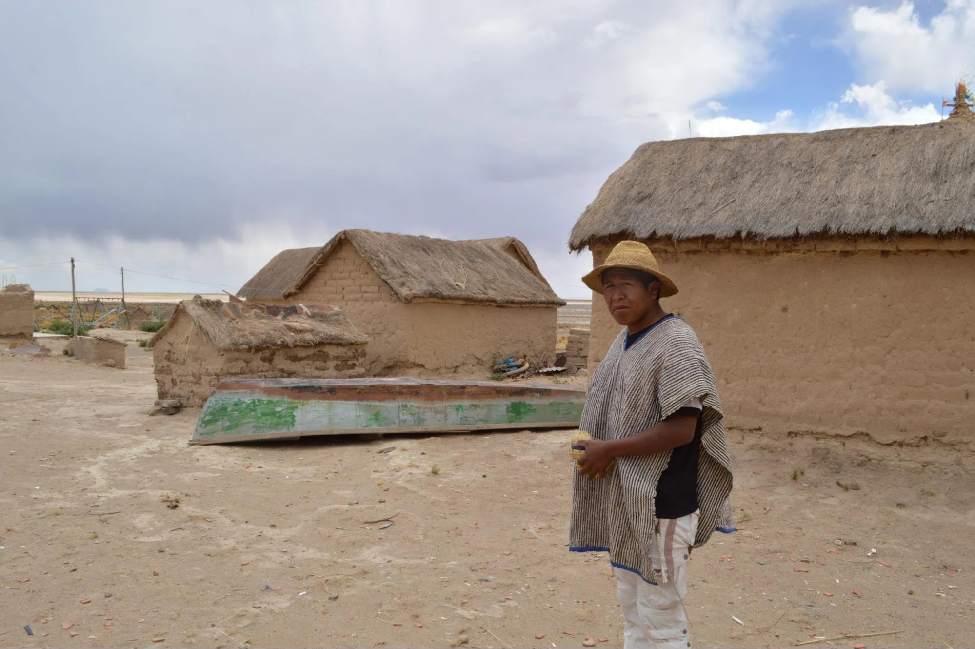 A man in a straw hat and striped poncho walking through a rural village of small adobe houses with thatched roofs.