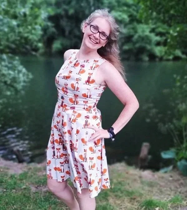 Amy Booth, a woman with blonde curly hair and glasses, smiling beside a river, wearing a white dress with a fox print. She looks happy, confident and approachable.