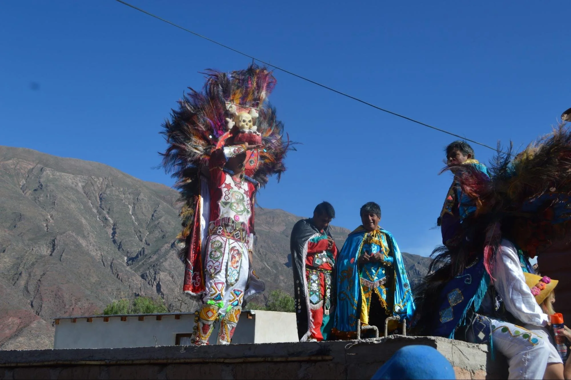 People in colourful traditional costume at an outdoor cultural festival, with mountains in the background and a beautiful, deep blue sky.