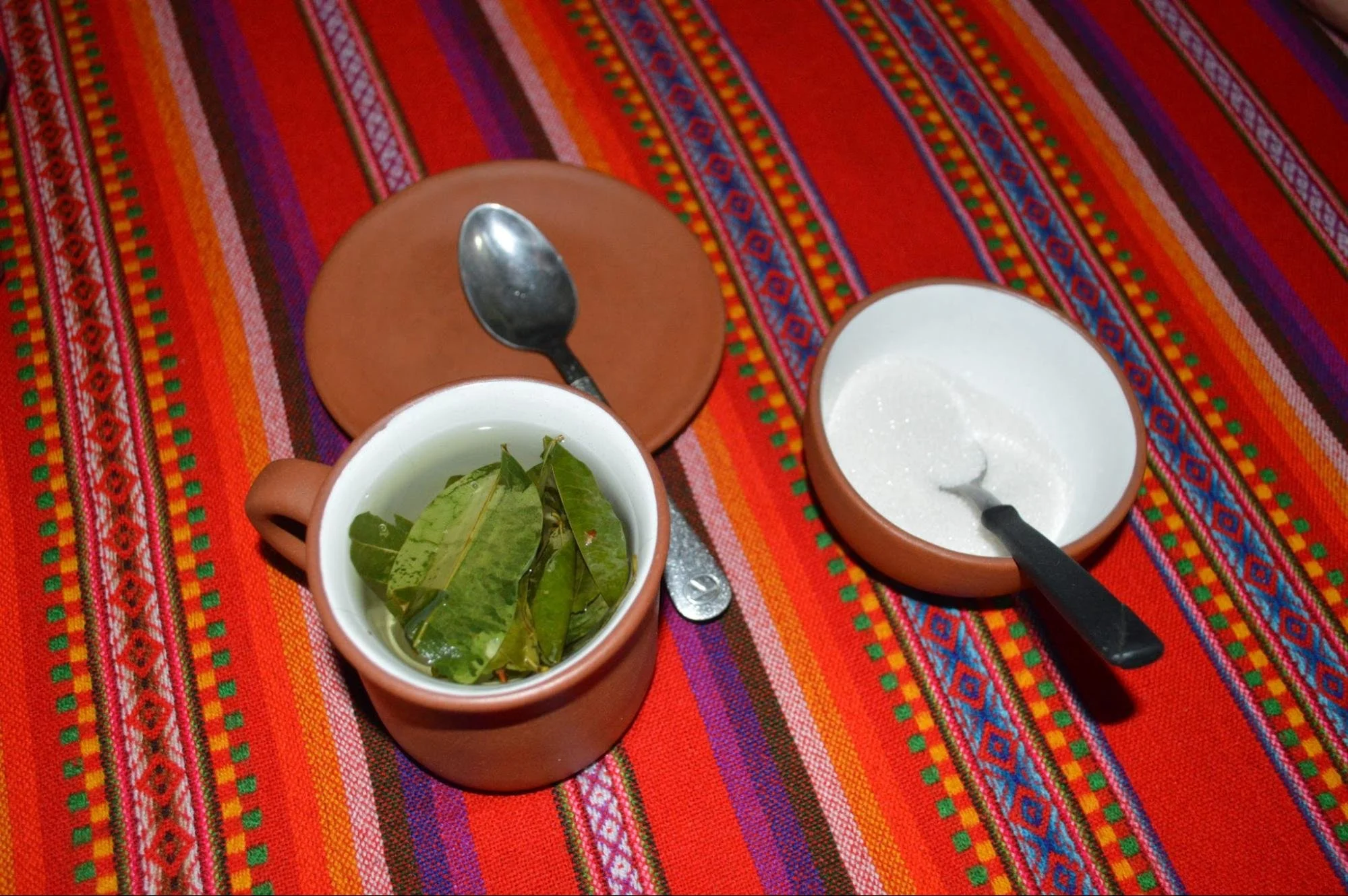 Three small terracotta bowls on colorful woven tablecloth, one with green tea leaves, one with sugar, and one with a spoon resting on a brown plate.