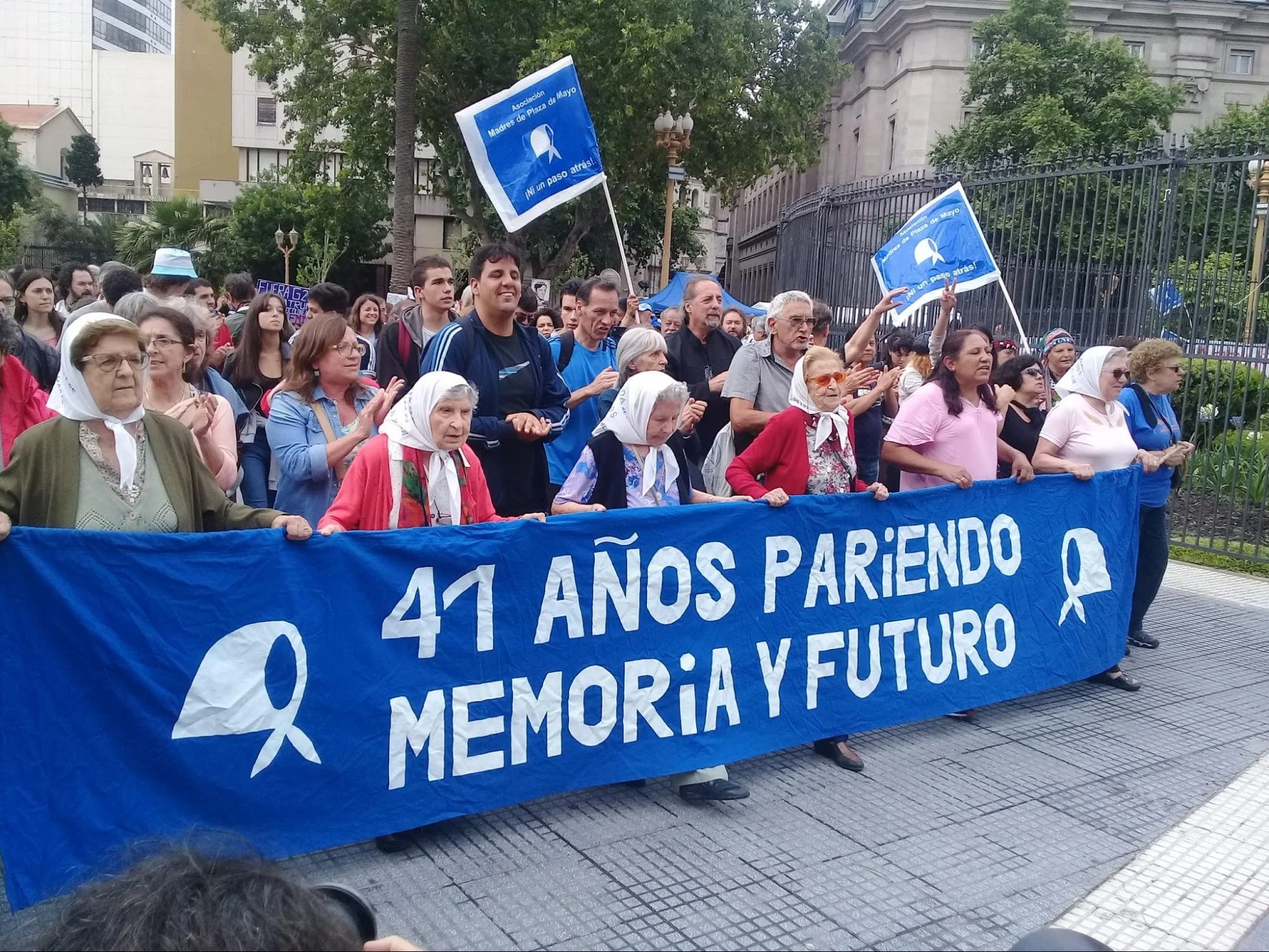 A daytime rally with banners referencing memory and the disappeared, attended by elderly women in headscarves and families, outside a gated building.