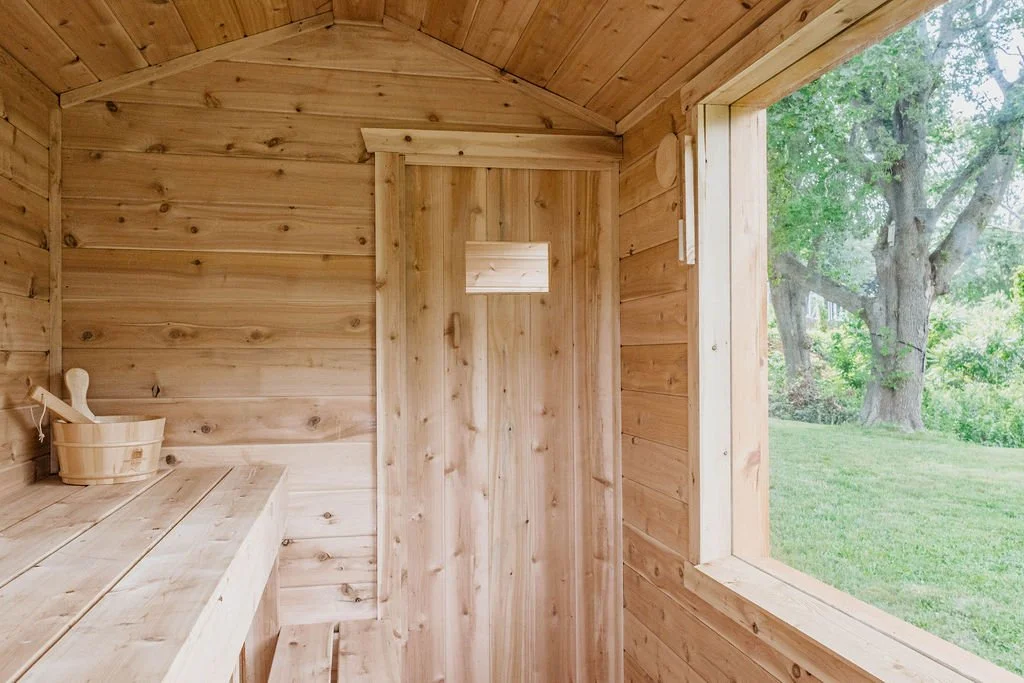 Inside a small wooden sauna with a window overlooking a green yard and large trees.