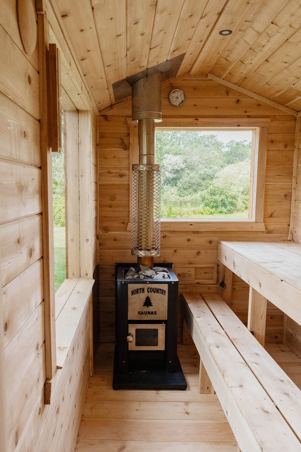 Interior of a wooden sauna with a wood stove, benches, a window with a view of trees, and a thermometer on the ceiling.