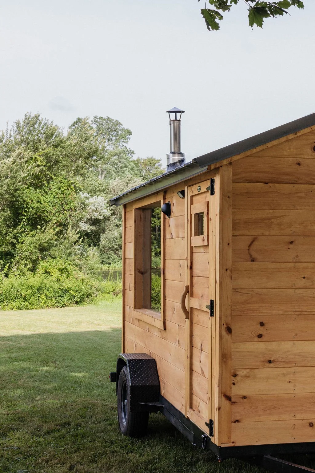 A small wooden mobile cabin or tiny house on wheels with an open window and a door, situated on a grassy area with trees in the background, and a chimney on the roof.