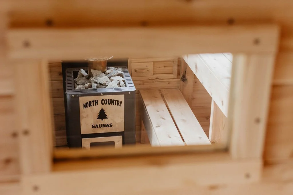 View of a sauna room through a small window, featuring a wood interior, a bench, and a heater with rocks labeled 'North Country Saunas'.