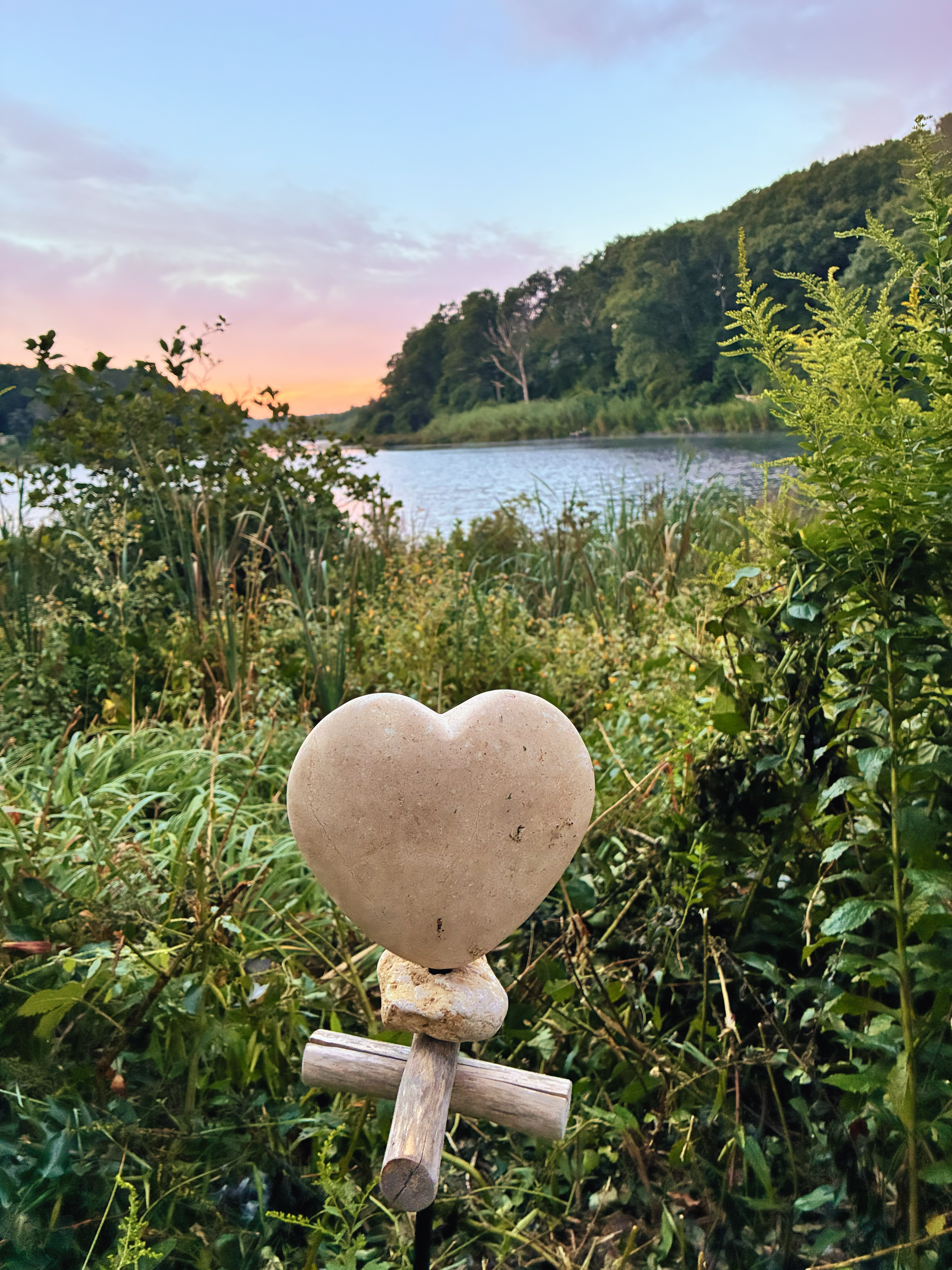 A heart-shaped stone sculpture on a T-shaped wooden base in a natural outdoor setting, with greenery and a river in the background during sunset.