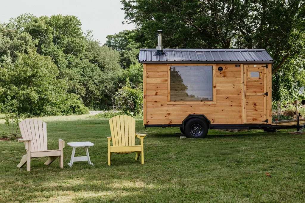 A small wooden tiny house on wheels with a black metal roof, large window, and door. Two Adirondack chairs and a small table sit on the grass nearby, with trees and greenery in the background.