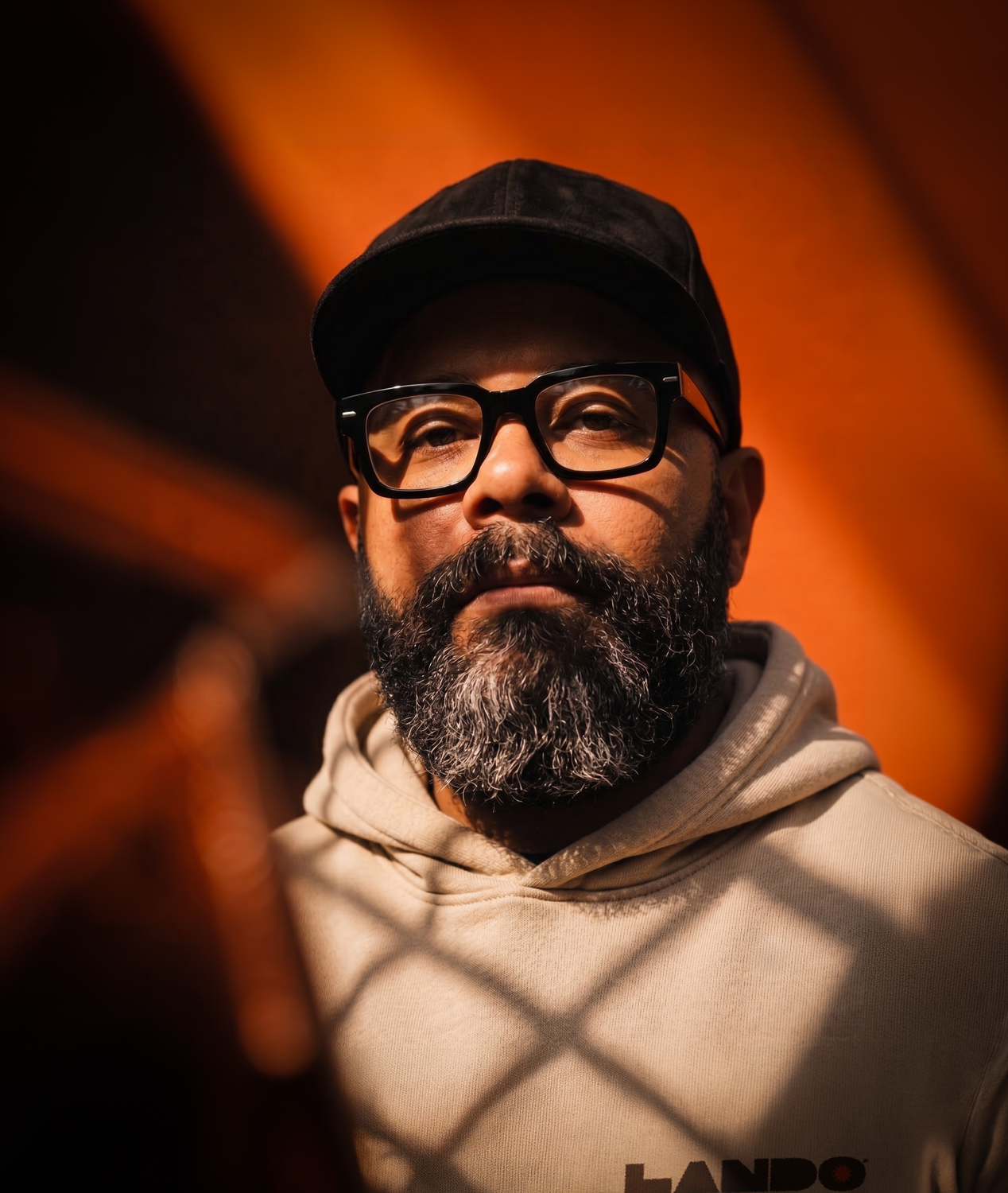 A close-up of a man with glasses, a black cap, and a beard, looking through a chain-link fence indoors with warm lighting.