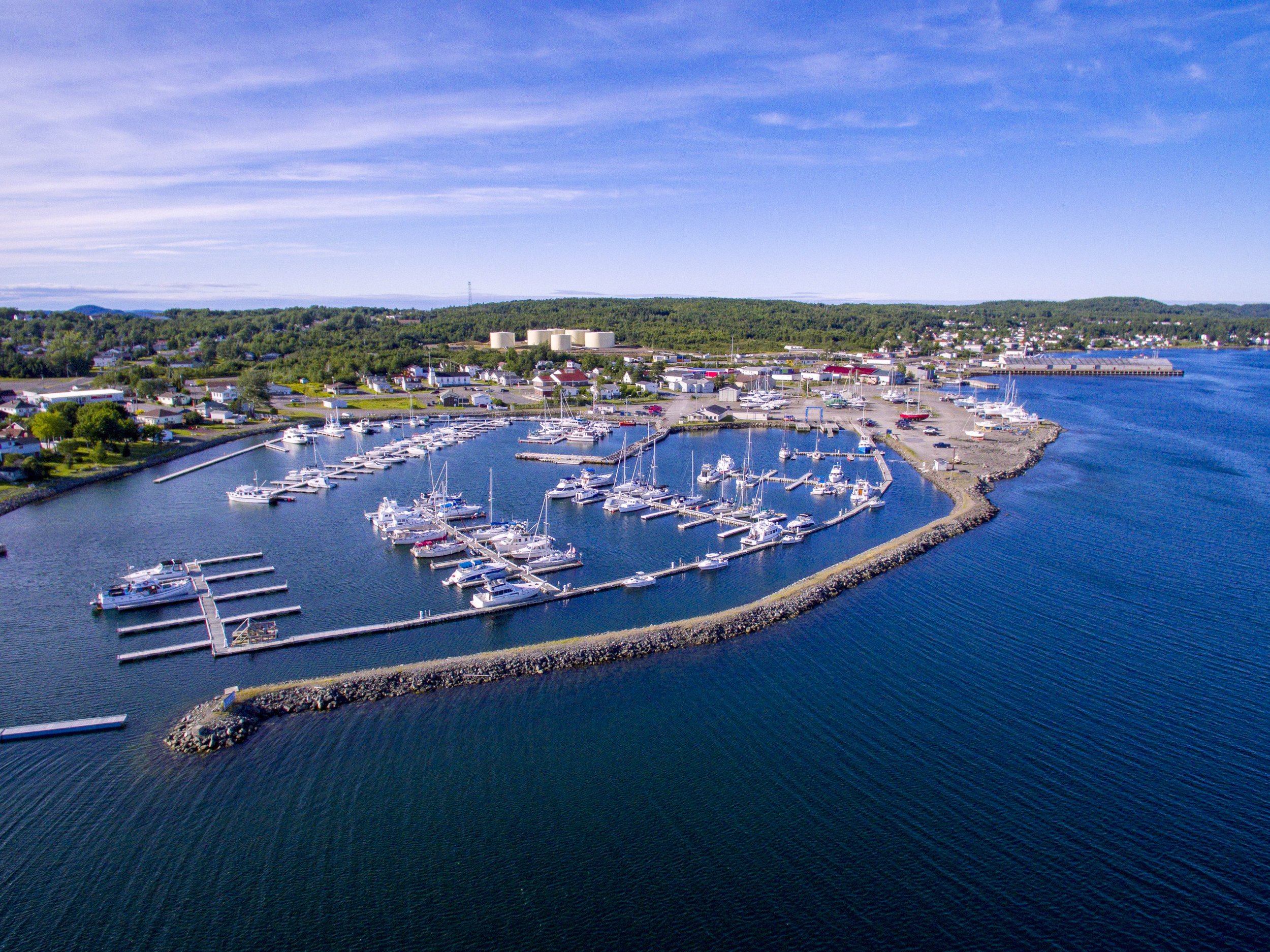 Aerial view of a marina with numerous boats docked, surrounded by water, with a small coastal town and green hills in the background under a blue sky with scattered clouds.