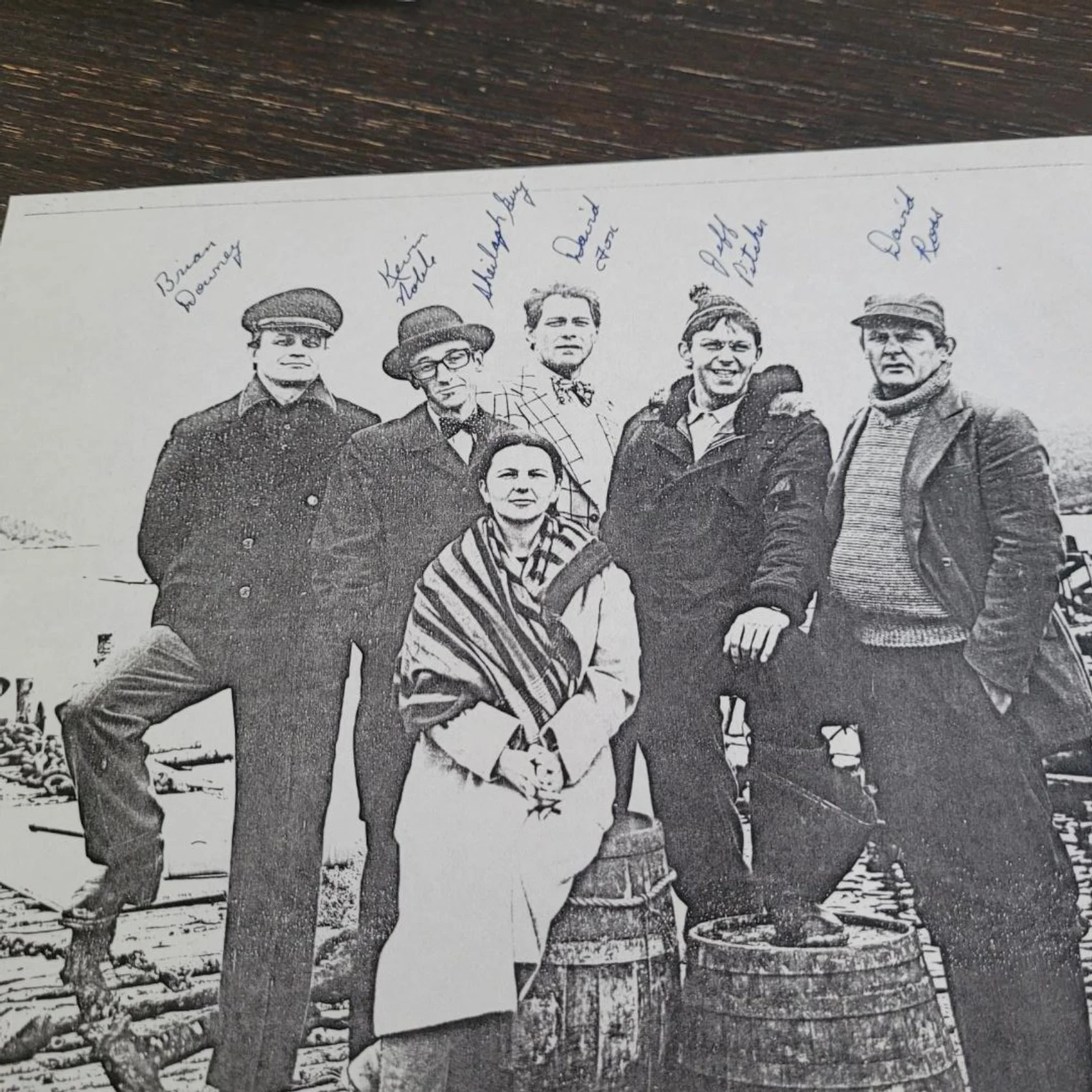 Black and white photograph of seven people standing outdoors in front of wooden barrels, with handwritten labels above each person, indicating their names.
