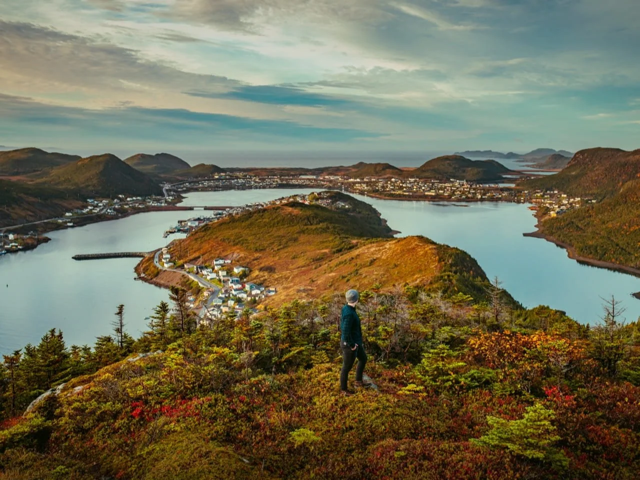 A person in outdoor clothing and a beanie standing on a hill overlooking a winding river, a town with numerous buildings, and surrounding mountains at sunset.