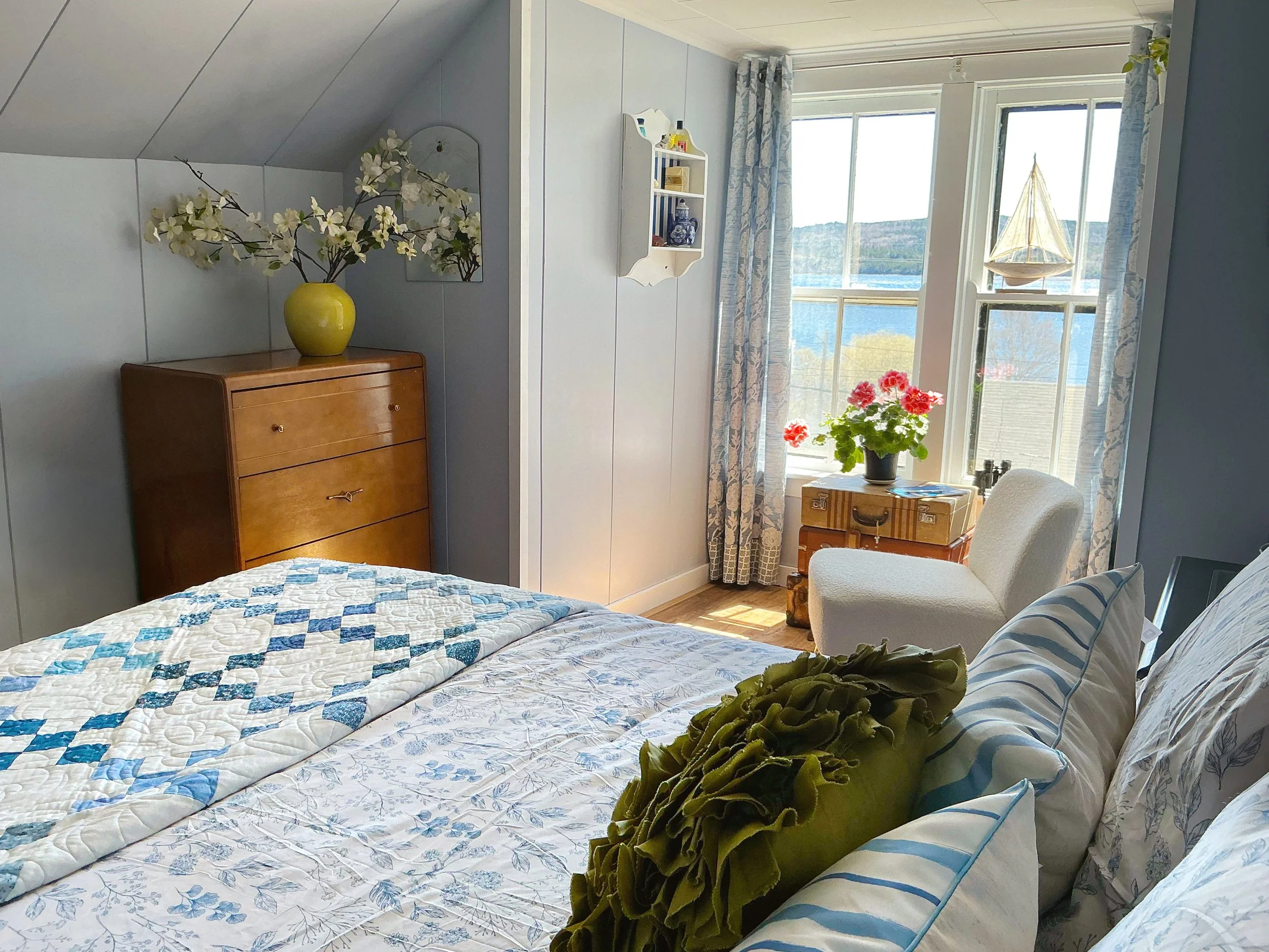 A bedroom with a bed covered in a quilt with blue and white patterns. There is a green plush toy on the bed, a white and blue pillow, and a window with curtains. Outside the window, a view of water and hills is visible. A wooden dresser with a yellow vase containing white flowers is in the corner, and a mirror behind it. A small white shelf with decorative items hangs on the wall near the window. A table with a potted plant with red flowers is nearby.