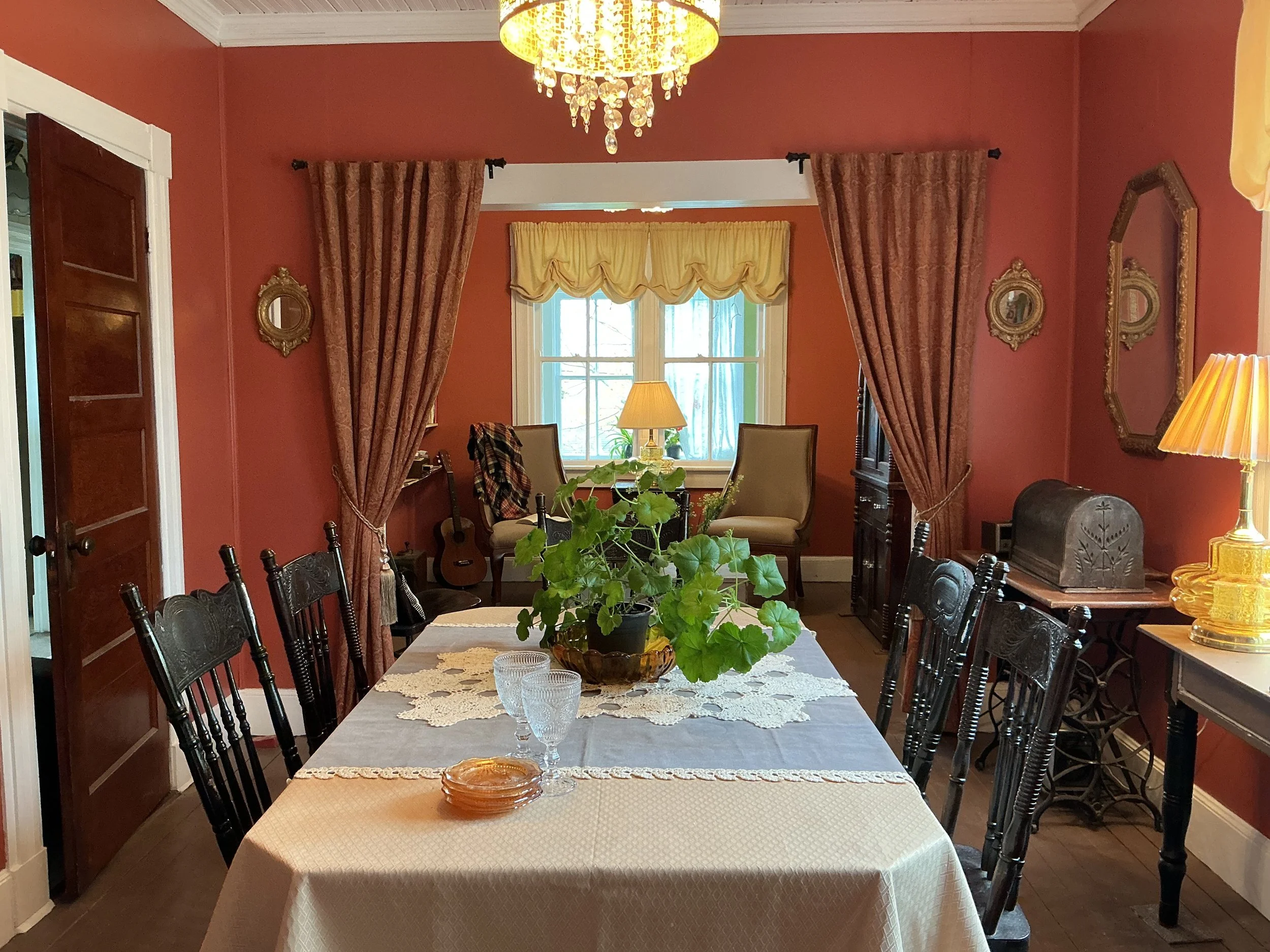 A dining room with a white tablecloth on the table, surrounded by black wooden chairs. There are green plants and glassware on the table. The room features red walls, golden framed mirrors, curtains, and a chandelier hanging from the ceiling. There are windows with yellow and white drapes, and various furniture including a lamp and a guitar in the background.