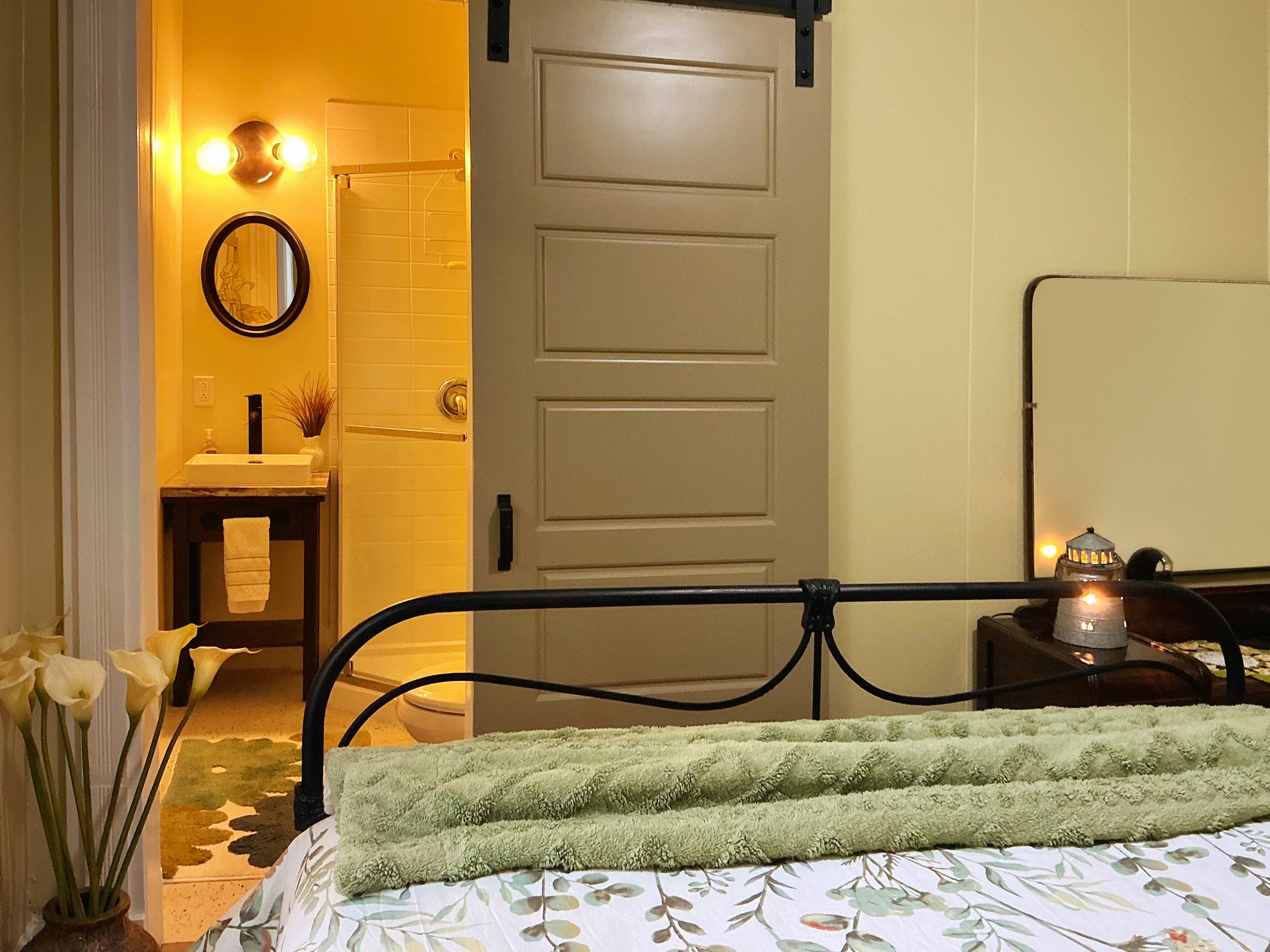 Bedroom view with plants and flowers in the foreground, a bed with a floral bedspread, and a green towel folded on it; in the background, a view into a bathroom with a mirror, sink, and shower area.