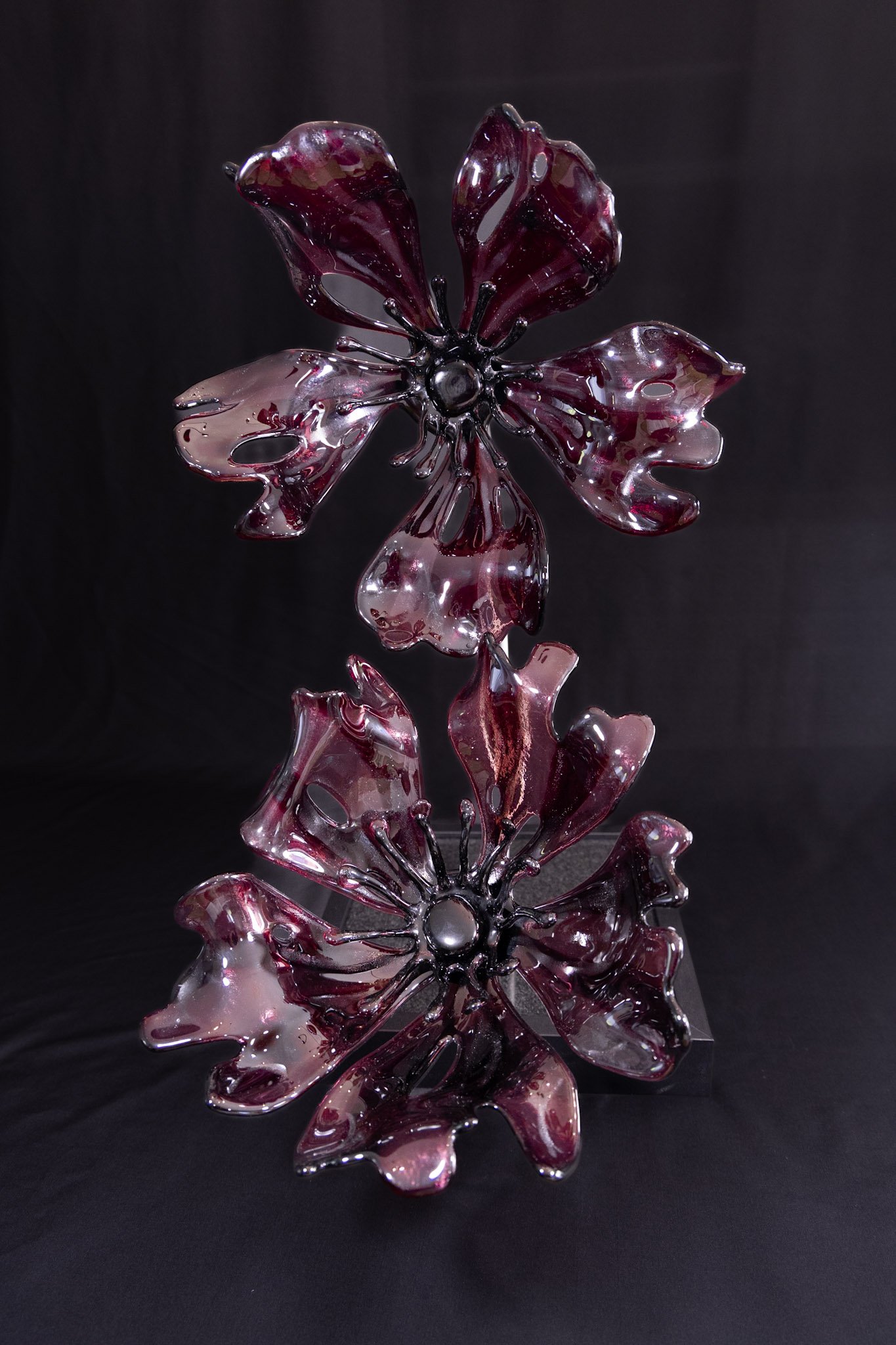 Decorative glass sculpture shaped like a flower with dark red and pink petals, reflected on a glossy black surface against a black background.