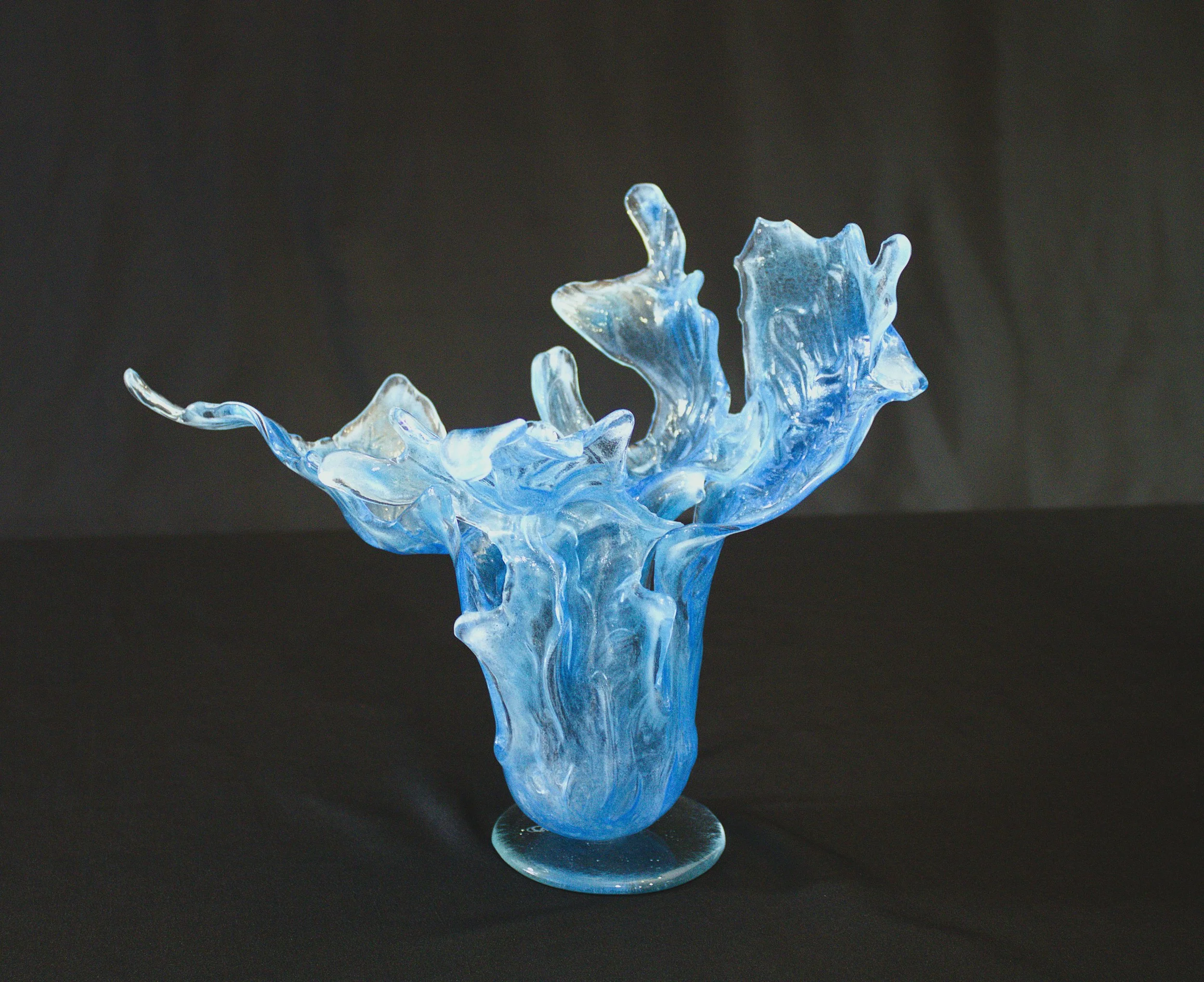 A glass sculpture resembling a splash of water, crafted from clear and blue-tinted glass, mounted on a round glass base against a black background.