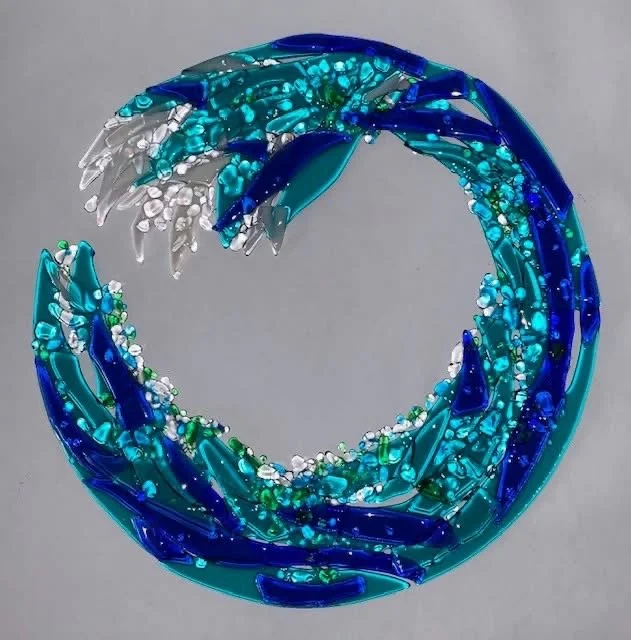 A decorative art piece of a circular wave made of blue, teal, white, and clear glass fragments on a gray background.