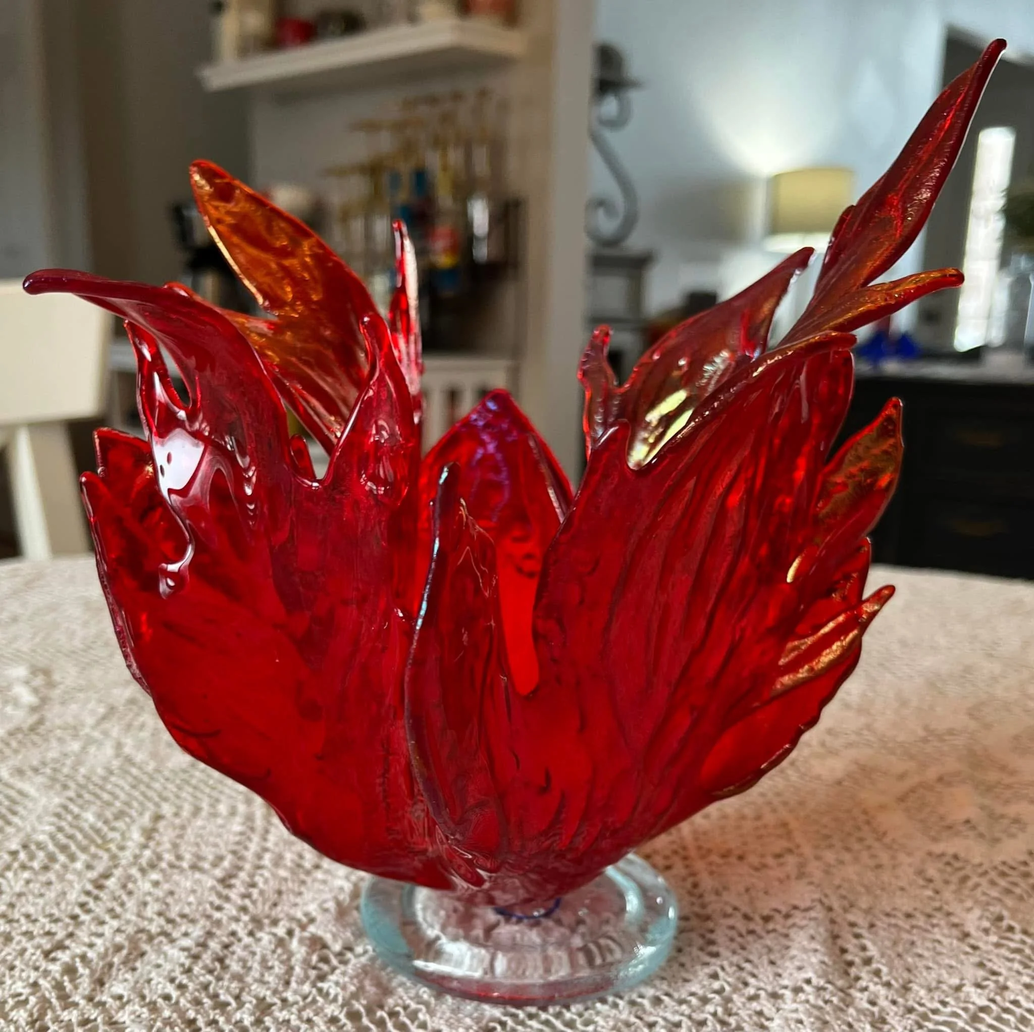Close-up of a red glass bowl with intricate patterns, placed on a beige carpet in a room with furniture and a lamp in the background.