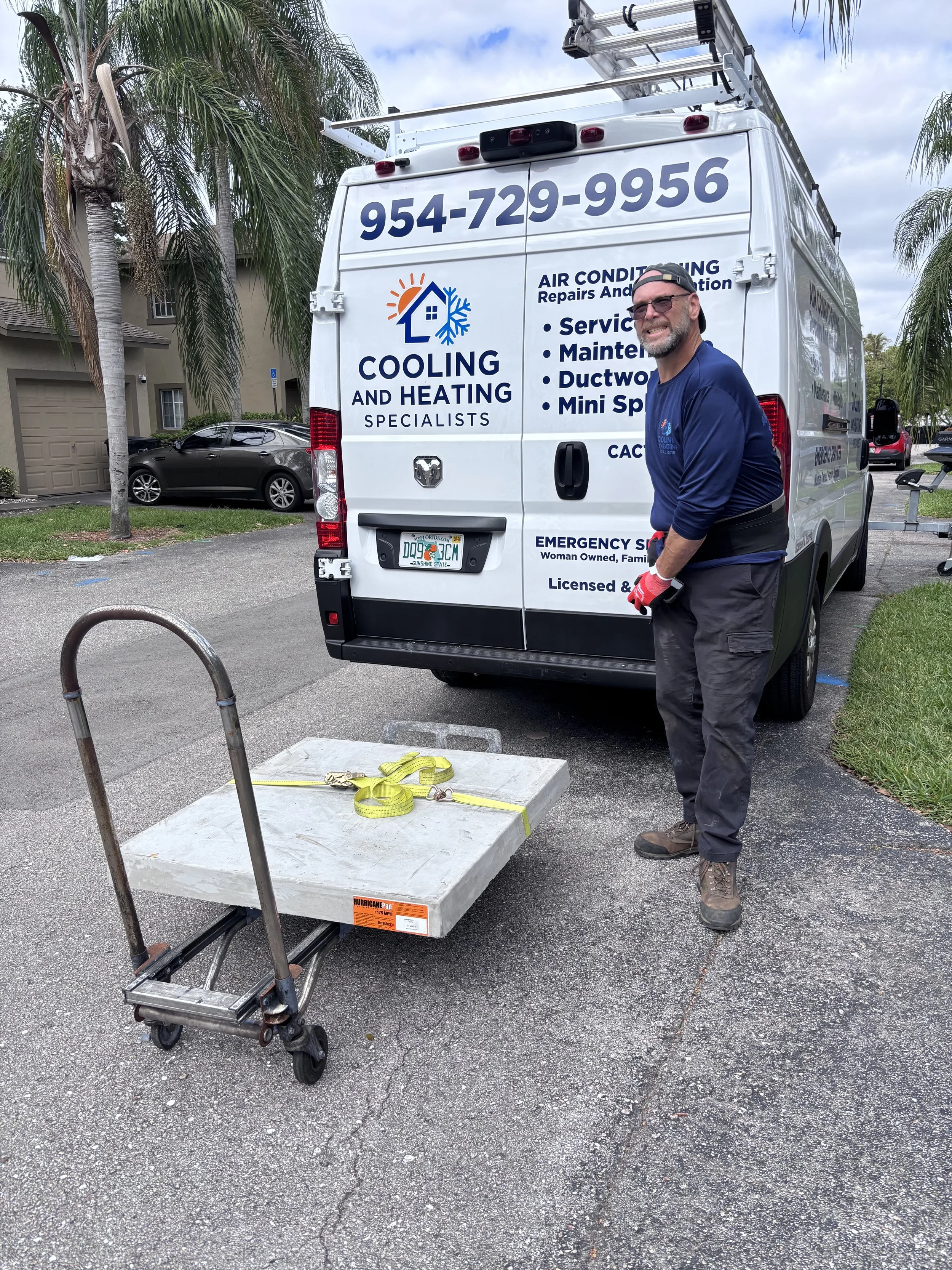 A man stands next to a white van with phone number and company logo for cooling and heating services. There is a cart and a large concrete slab on the ground in front of him. The background has some houses, a parked car, and palm trees.
