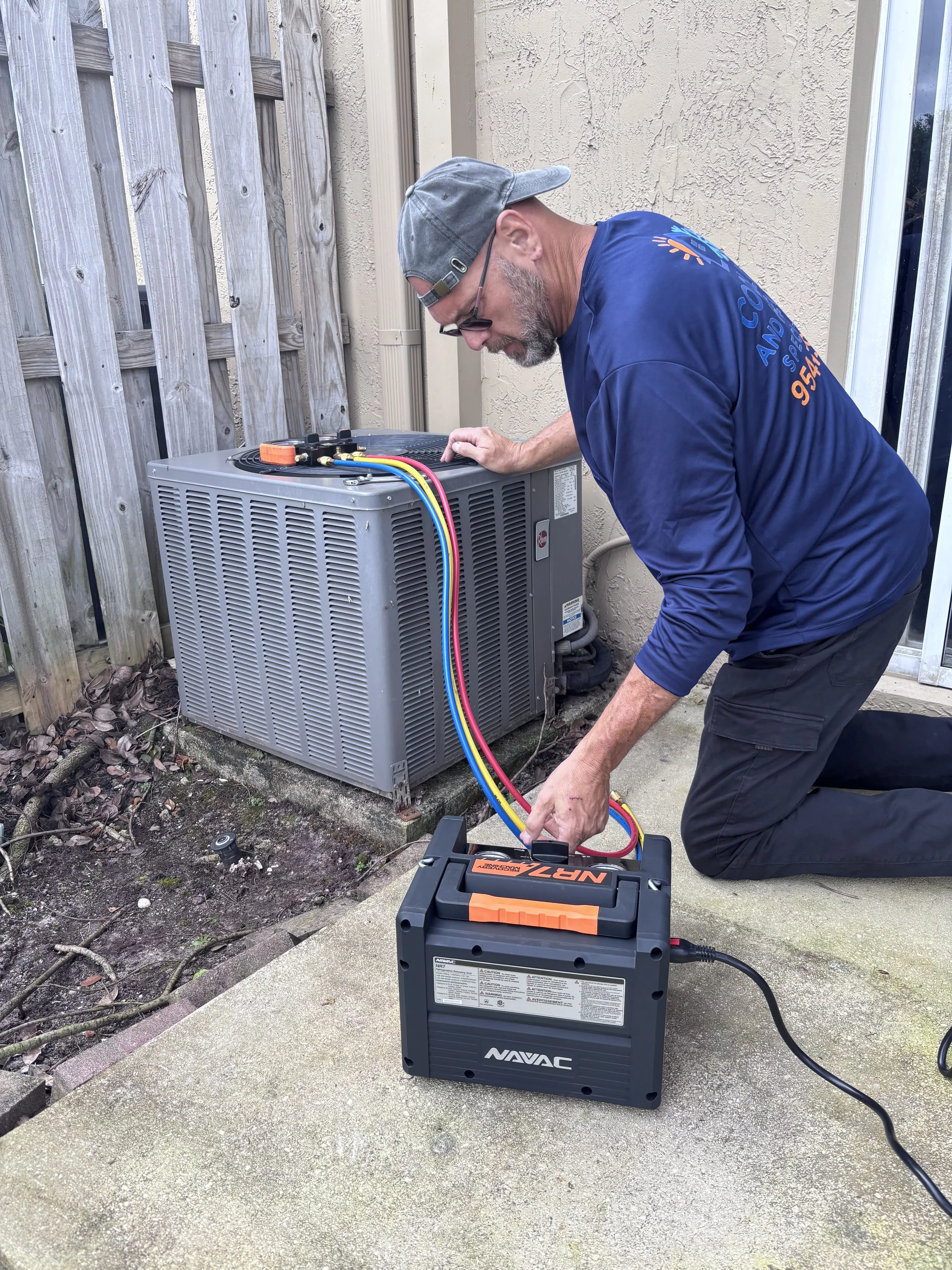 A man kneeling outdoors on a concrete patio, working on an air conditioning unit with jumper cables and a portable battery.