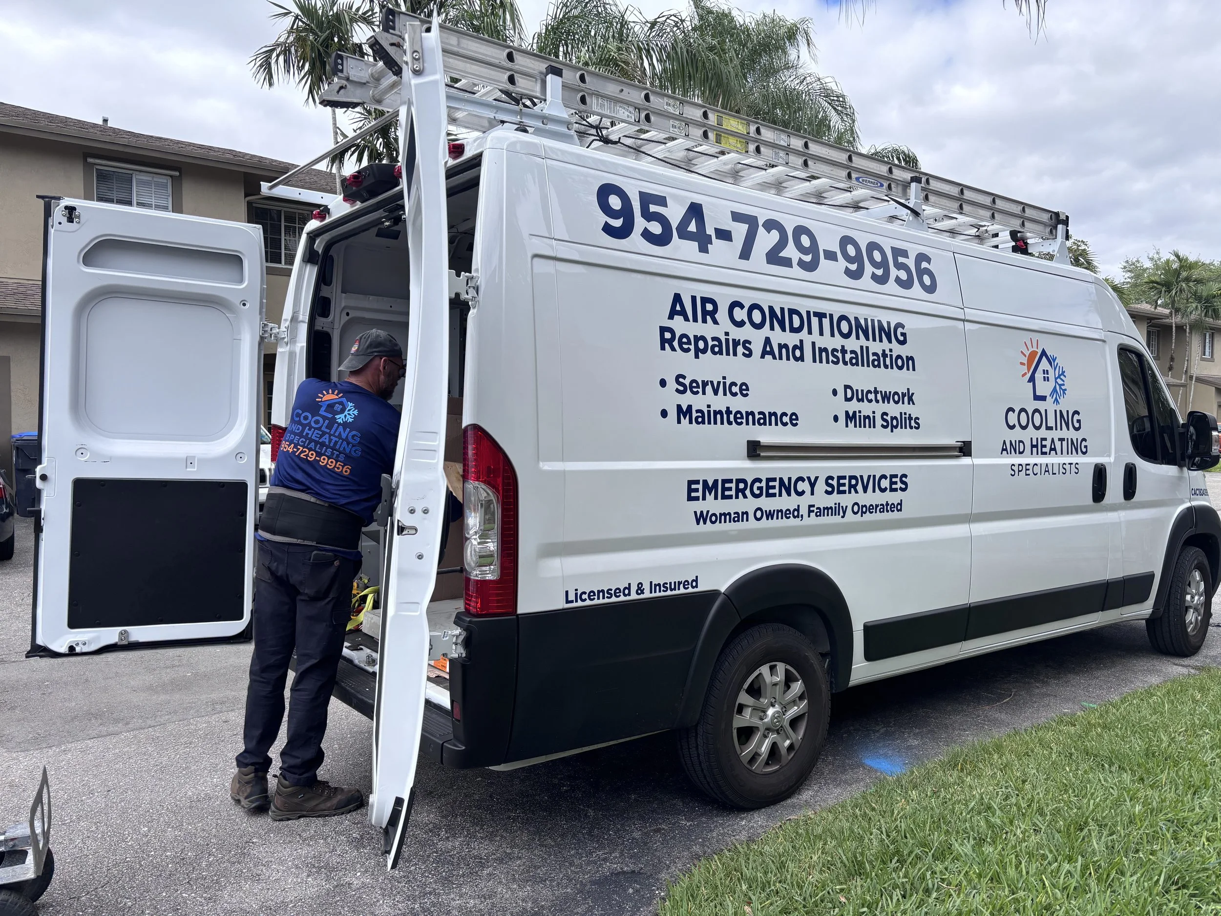 A white service van for cooling and heating specialists with a ladder on top, open side door revealing tools, parked on a residential street with a man in uniform working nearby